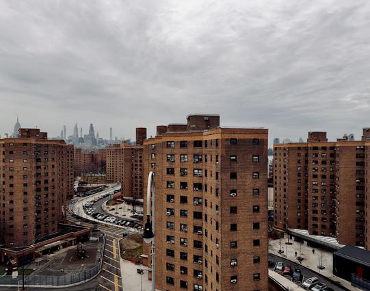 A color photograph in square aspect ratio. An elevated, high-angle view looks north over a dense complex of multi-story, red-brick housing buildings in the Lower East Side neighborhood of Manhattan. The foreground and middle ground are dominated by several identical, cruciform-shaped apartment towers with many small windows and protruding air conditioning units. Between the buildings, a curved asphalt road is lined with parked cars, and a small playground with a green slide is visible at the bottom left. The sky is a heavy, uniform grey with thick cloud cover, casting a flat, somber light over the urban landscape. In the far distance, the midtown Manhattan skyline rises as a jagged silhouette against the overcast horizon. Iconic skyscrapers, including the Empire State Building and several slender "pencil towers," are visible, though somewhat hazy due to the distance and weather conditions. A glimpse of the East River is visible to the right between the apartment blocks.