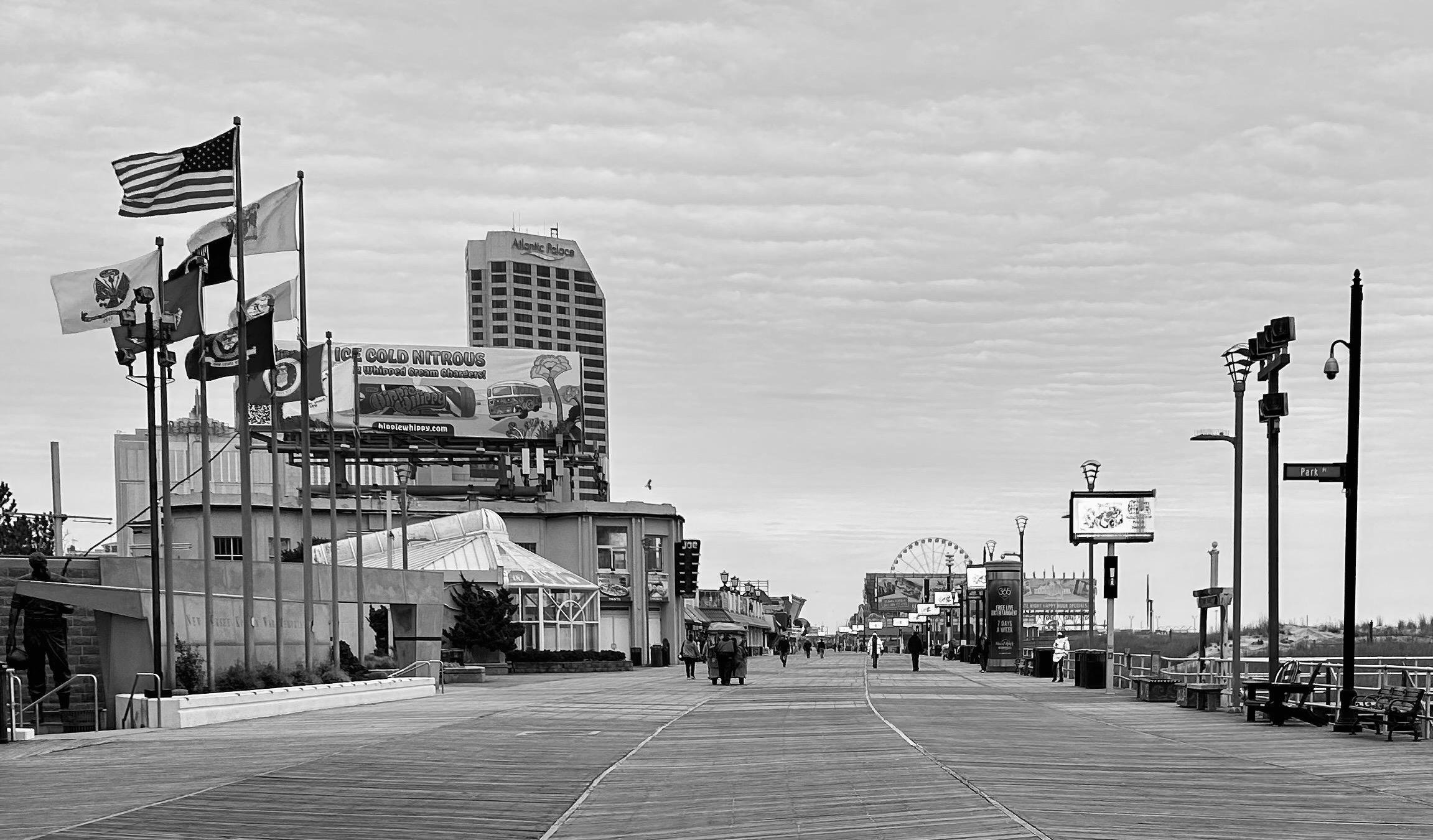 A noir, wide-angle photograph in landscape orientation that captures a vanishing-point perspective of the Atlantic City Boardwalk on a quiet, overcast winter day. In the foreground to the left stands the New Jersey Korean War Memorial, marked by a cluster of tall flagpoles flying the American flag alongside various military and state banners. Beside the memorial, a large billboard advertises "Ice Cold Nitrous," and the glass-enclosed pavilion of a nearby building sits nestled behind the boardwalk's edge. The weathered wooden planks of the boardwalk feature distinct walking paths created by darker, weathered lines in the wood, stretching deep into the distance where a few scattered pedestrians and a rolling chair stroll under a pale, streaked sky. In the mid-ground and background, the skyline of the city rises against the horizon. The tall, rectangular Atlantic Palace building dominates the left-center view, while a Ferris wheel is visible further down the boards to the right. The right side of the frame is lined with modern street lamps, benches, and a Park Place street sign, bordering a sandy area that transitions toward the beach. The overall atmosphere is serene and slightly desolate, capturing the off-season stillness.