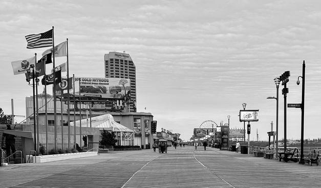 A noir, wide-angle photograph in landscape orientation that captures a vanishing-point perspective of the Atlantic City Boardwalk on a quiet, overcast winter day. In the foreground to the left stands the New Jersey Korean War Memorial, marked by a cluster of tall flagpoles flying the American flag alongside various military and state banners. Beside the memorial, a large billboard advertises "Ice Cold Nitrous," and the glass-enclosed pavilion of a nearby building sits nestled behind the boardwalk's edge. The weathered wooden planks of the boardwalk feature distinct walking paths created by darker, weathered lines in the wood, stretching deep into the distance where a few scattered pedestrians and a rolling chair stroll under a pale, streaked sky. In the mid-ground and background, the skyline of the city rises against the horizon. The tall, rectangular Atlantic Palace building dominates the left-center view, while a Ferris wheel is visible further down the boards to the right. The right side of the frame is lined with modern street lamps, benches, and a Park Place street sign, bordering a sandy area that transitions toward the beach. The overall atmosphere is serene and slightly desolate, capturing the off-season stillness.
