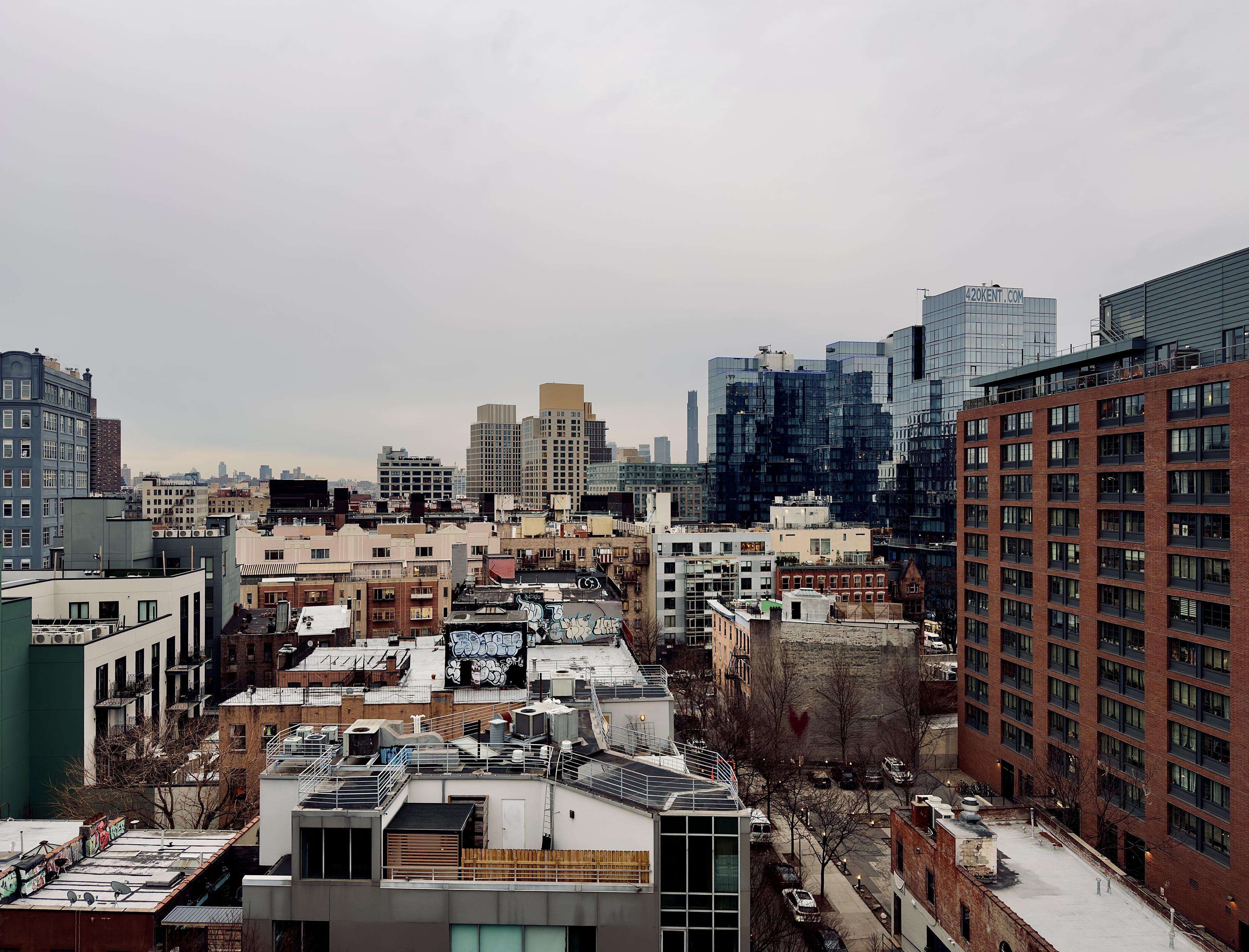 A color, wide-angle photograph that captures an elevated view of the dense urban landscape of South Williamsburg, Brooklyn, under a heavy, overcast gray sky. In the foreground and midground, a mix of low-to-mid-rise residential and commercial buildings creates a textured skyline of rooftops. These structures feature a variety of architectural styles, from older brick buildings with visible fire escapes and graffiti-covered water towers to modern white and gray developments with clean lines and glass balconies. To the right, a large, modern red-brick apartment complex dominates the frame, standing adjacent to a series of sleek, blue-glass high-rises. One of these glass towers prominently displays the "420KENT.COM" signage at its peak. In the far distance, the silhouette of skyscrapers is visible through the hazy atmosphere. Below the rooftops, a narrow street is lined with bare winter trees and a few parked cars. The overall mood is quiet and industrial.