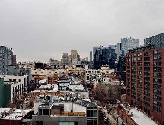A color, wide-angle photograph that captures an elevated view of the dense urban landscape of South Williamsburg, Brooklyn, under a heavy, overcast gray sky. In the foreground and midground, a mix of low-to-mid-rise residential and commercial buildings creates a textured skyline of rooftops. These structures feature a variety of architectural styles, from older brick buildings with visible fire escapes and graffiti-covered water towers to modern white and gray developments with clean lines and glass balconies. To the right, a large, modern red-brick apartment complex dominates the frame, standing adjacent to a series of sleek, blue-glass high-rises. One of these glass towers prominently displays the "420KENT.COM" signage at its peak. In the far distance, the silhouette of skyscrapers is visible through the hazy atmosphere. Below the rooftops, a narrow street is lined with bare winter trees and a few parked cars. The overall mood is quiet and industrial.
