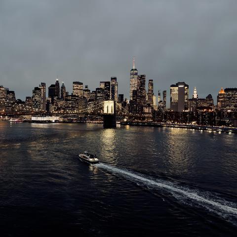 A color, wide-angle photograph in square aspect ratio that captures the Lower Manhattan skyline and the Brooklyn Bridge at twilight under a heavy, overcast winter sky. In the foreground, the dark, rippling waters of the East River are interrupted by a white ferry traveling toward the left, leaving a long, foaming white wake that angles toward the bottom right corner. The Brooklyn Bridge stretches across the mid-ground, its iconic stone arches illuminated by warm yellow lights that reflect off the water's surface. The background is dominated by the dense thicket of skyscrapers in the Financial District, glowing with thousands of interior lights against the deep grey atmosphere. One World Trade Center stands as the tallest spire, capped with a red, white and blue light. Other notable landmarks, including the Verizon Building and various Art Deco towers, create a jagged, glittering silhouette. The overall mood is moody and cinematic, defined by the contrast between the dark, cool tones of the sky and river against the warm, golden glow of the city's evening lights.