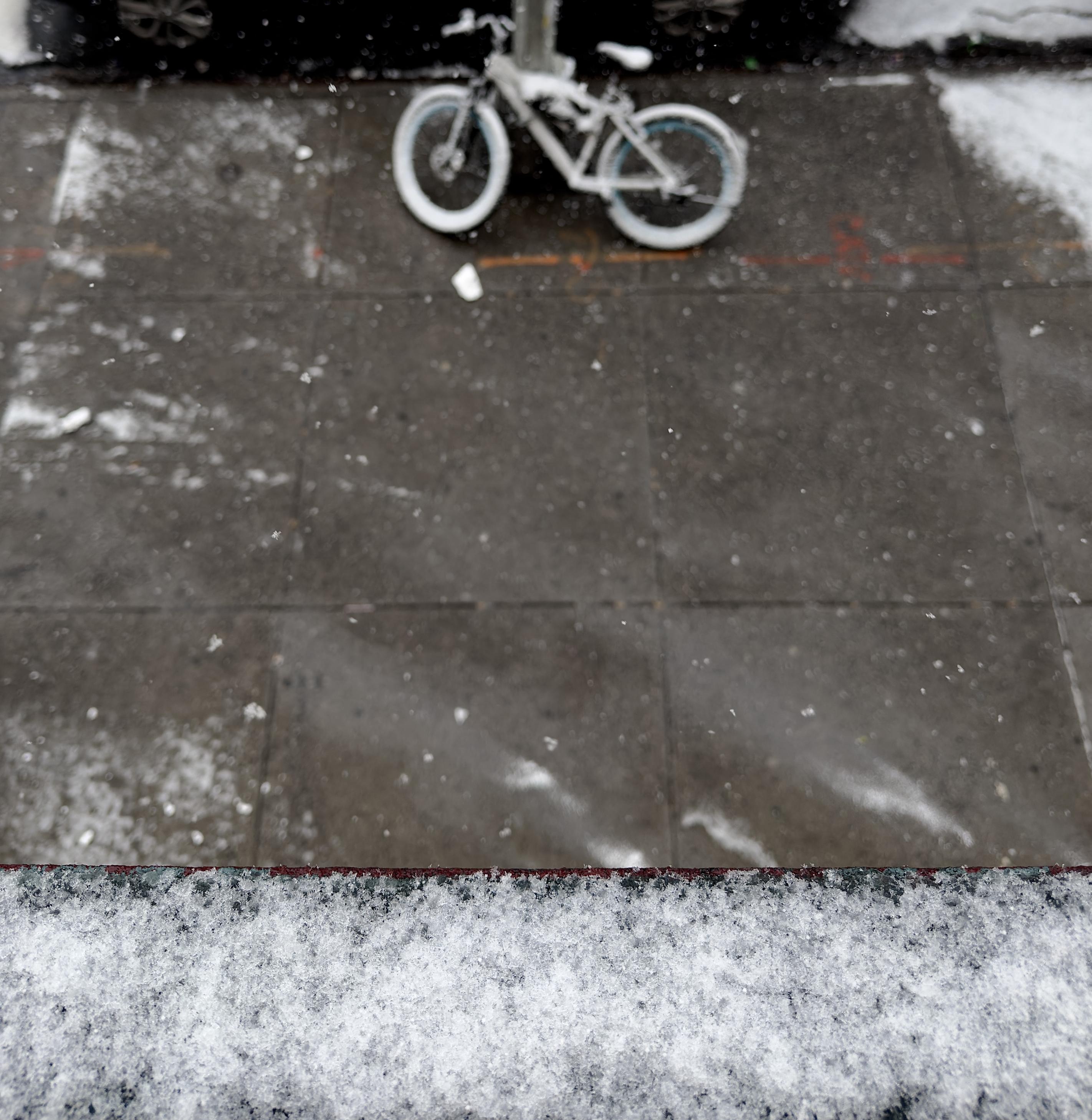 A color photograph in square aspect ratio. An overhead shot captures the beginnings of a light dusting of snow beginning to coat a Manhattan sidewalk. In the upper center, a bicycle with white-walled tires is parked against a metal pole, its seat and frame lightly dusted with white. The dark, damp concrete of the sidewalk below is speckled with individual snowflakes and small, patchy accumulations, while a prominent, icy-white layer of snow clings to a ledge in the immediate foreground. Beyond the bike, the edges of the street show thicker white buildup against a dark curb.