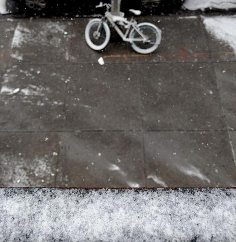A color photograph in square aspect ratio. An overhead shot captures the beginnings of a light dusting of snow beginning to coat a Manhattan sidewalk. In the upper center, a bicycle with white-walled tires is parked against a metal pole, its seat and frame lightly dusted with white. The dark, damp concrete of the sidewalk below is speckled with individual snowflakes and small, patchy accumulations, while a prominent, icy-white layer of snow clings to a ledge in the immediate foreground. Beyond the bike, the edges of the street show thicker white buildup against a dark curb.