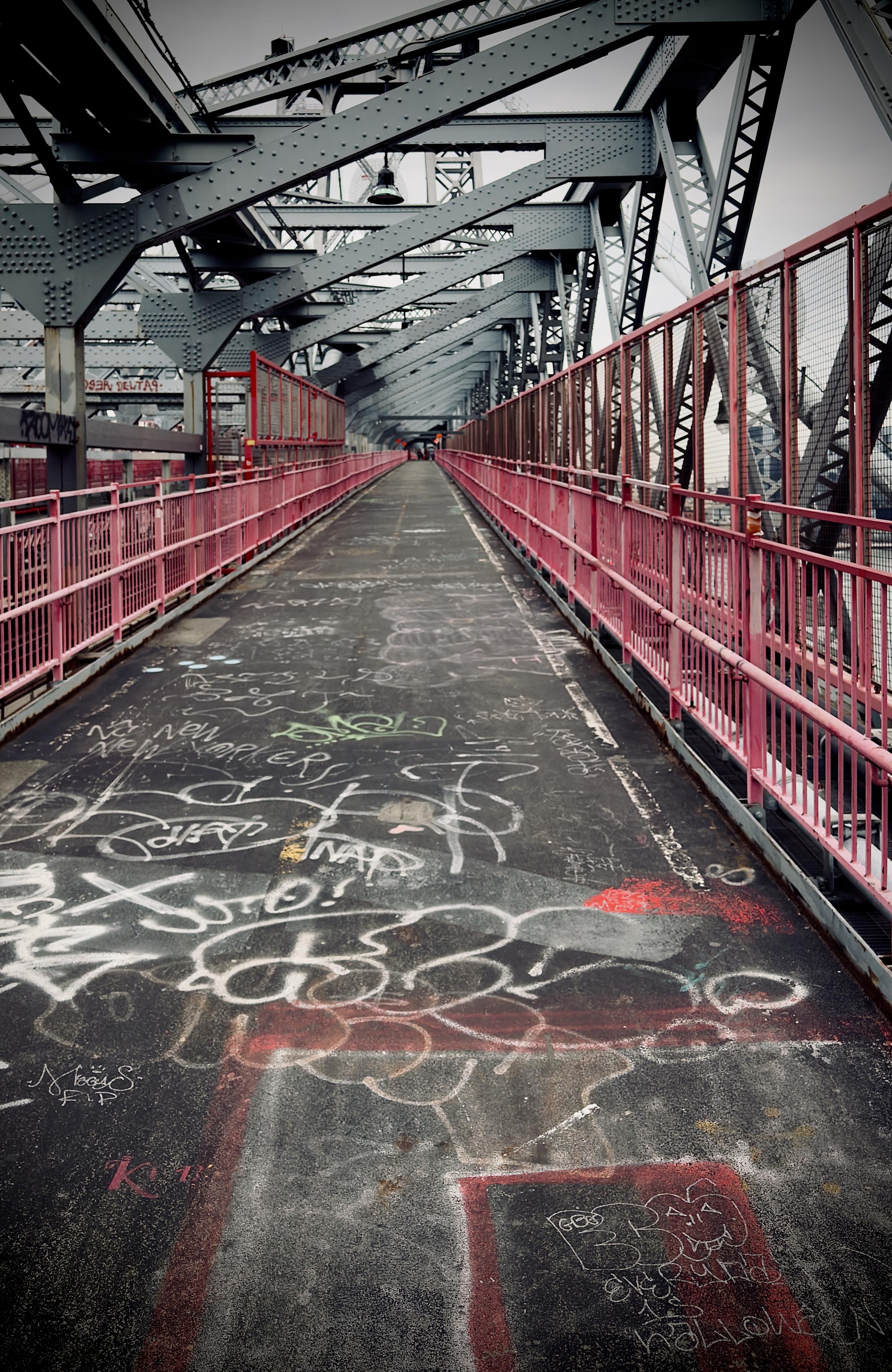 A color photograph in portrait orientation that captures the distinctive industrial character of the Williamsburg Bridge pedestrian walkway in New York City on an overcast afternoon. The scene is dominated by the bridge's massive pink-painted steel trusses and grey support beams that crisscross overhead, casting geometric shadows across the path. The walkway itself is paved in weathered asphalt, heavily layered with vibrant, overlapping graffiti tags and street art in shades of neon pink, yellow, and white. A chain-link fence runs along the side, offering a glimpse of the East River and the Brooklyn skyline through the steel mesh. The path appears slightly damp, highlighting the textures of the grit and colorful scrawl on the ground. To one side, the subway tracks are visible behind a heavy iron grate.