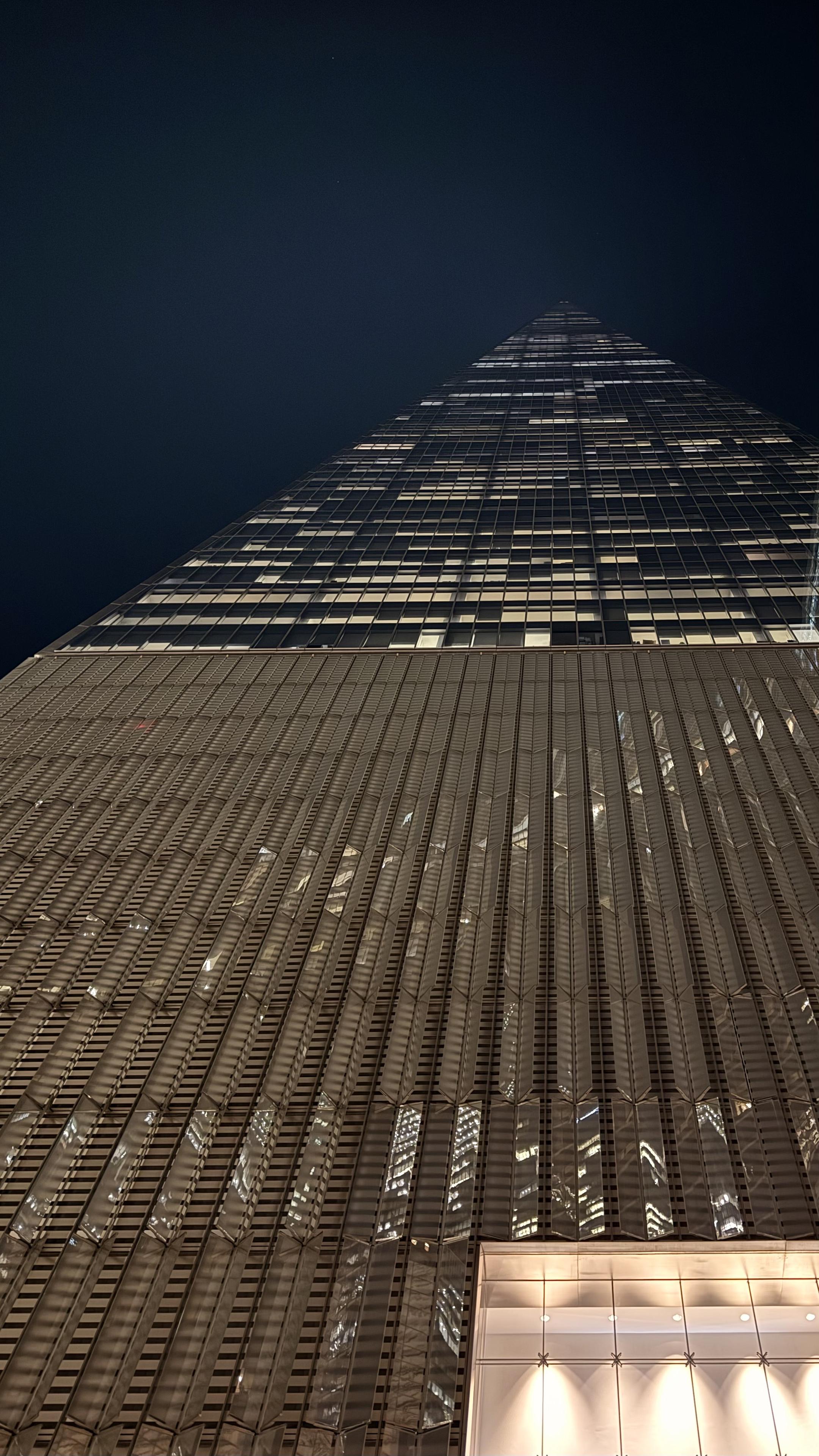 A color, low-angle photograph of the One World Trade Center tower in New York City taken at night. The composition looks sharply upward, emphasizing the immense height and tapering geometry of the skyscraper. The building dominates the frame against a completely black sky. The lower two-thirds of the image show the tower's base, illuminated entrance, and the initial tiers of its reflective glass, characterized by vertical fins and sharp architectural lines. These sections reflect distorted, bright lights from the interior and surrounding buildings, creating a textured, almost abstract pattern of golden light and shadow that runs along the glass surface. The upper third of the image is dedicated to the tower's ascent, where the structure narrows toward its pinpoint shaped apex. In this section, the illuminated office windows create a striking grid pattern, with many windows displaying bright squares of light that stand out against the otherwise dark glass and hazy deep blue night sky.