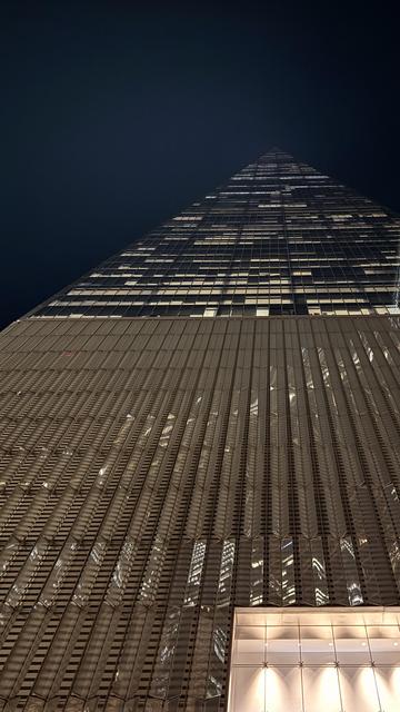 A color, low-angle photograph of the One World Trade Center tower in New York City taken at night. The composition looks sharply upward, emphasizing the immense height and tapering geometry of the skyscraper. The building dominates the frame against a completely black sky. The lower two-thirds of the image show the tower's base, illuminated entrance, and the initial tiers of its reflective glass, characterized by vertical fins and sharp architectural lines. These sections reflect distorted, bright lights from the interior and surrounding buildings, creating a textured, almost abstract pattern of golden light and shadow that runs along the glass surface. The upper third of the image is dedicated to the tower's ascent, where the structure narrows toward its pinpoint shaped apex. In this section, the illuminated office windows create a striking grid pattern, with many windows displaying bright squares of light that stand out against the otherwise dark glass and hazy deep blue night sky.