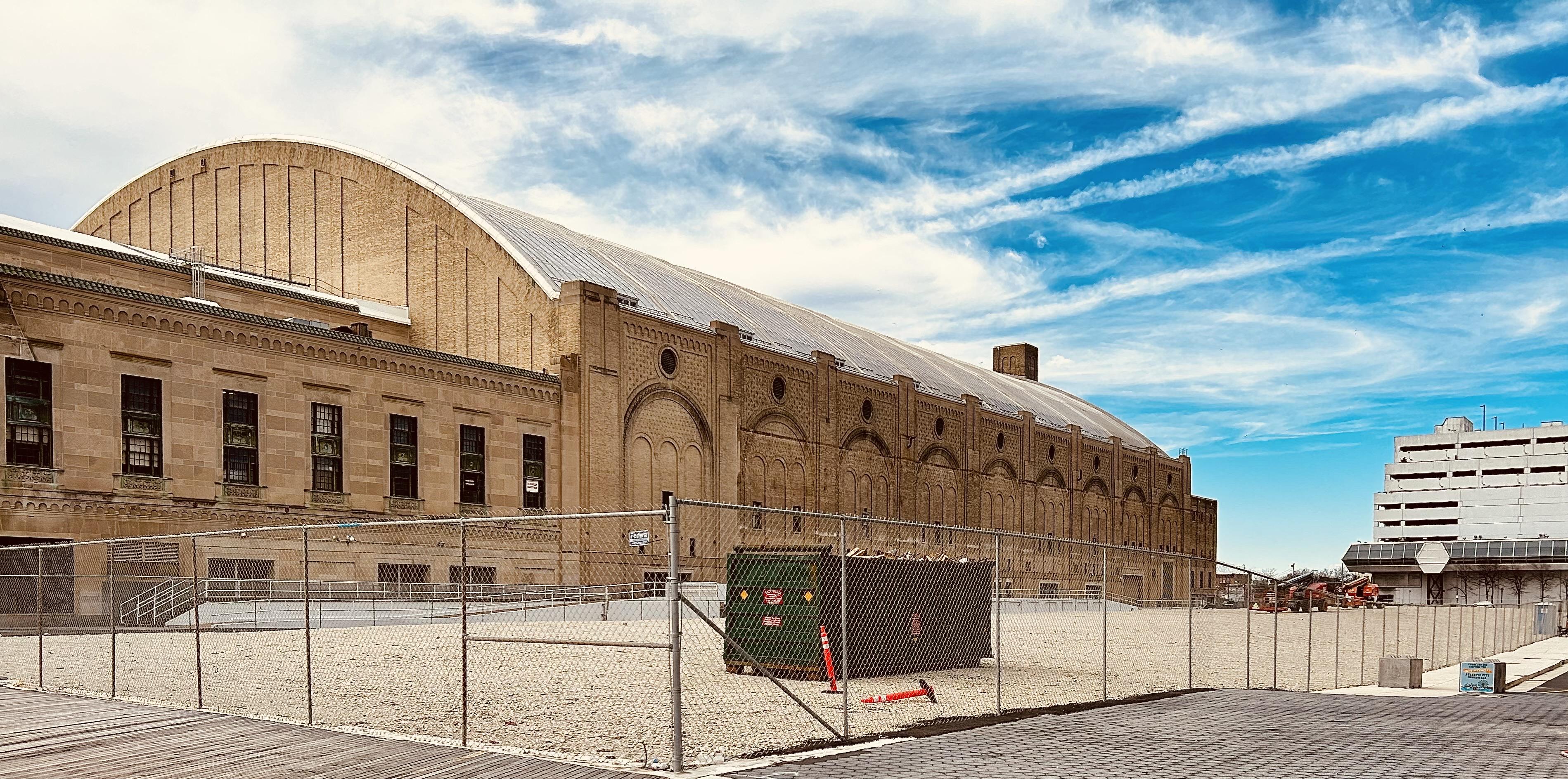 A color, wide-angle photograph in landscape orientation that captures the vacant lot where the Trump Plaza Hotel and Casino once stood in Atlantic City, New Jersey. The lot is a wide, flat expanse covered in light-colored gravel and surrounded by a tall chain-link fence. Near the center of the site, a large green industrial dumpster and a few orange traffic markers sit on the gravel. In the background, a white multi-story parking garage remains standing to the right, while a construction crane is visible further in the distance. Dominating the left side of the frame is the historic Jim Whelan Boardwalk Hall, a massive limestone and brick building featuring a soaring, arched roofline and classical architectural details. Its weathered tan facade and rows of dark windows contrast with the bright, streaky deep blue sky filled with wispy white clouds. The foreground consists of the boardwalk area leading up to the fenced perimeter of the empty site. There are no people in the scene.