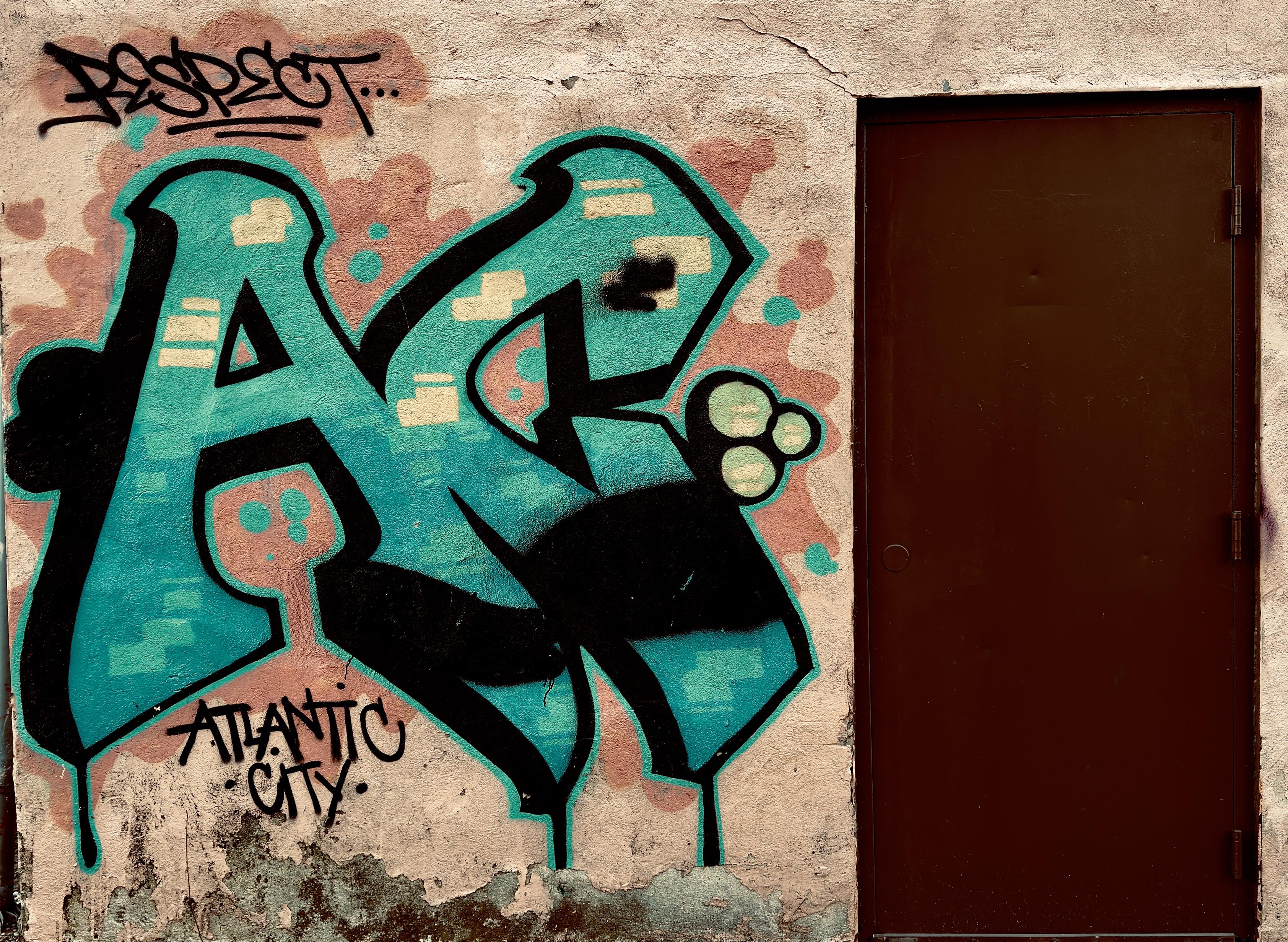 A color, medium-angle photograph taken outside of a liquor store in Atlantic City, New Jersey. The focal point of the image is a vibrant piece of graffiti street art that is spray-painted onto a weathered, textured beige exterior wall next to a solid, dark brown metal door. The central focus is a large, stylized piece of lettering in a bright teal and turquoise hue, outlined in bold black. The letters "AC” feature blocky, geometric shapes with internal highlights of pale yellow and black shading to create a three-dimensional effect. Above the main letters, a black tag reads "RESPECT..." in a classic graffiti script with two underlines, while below the letters, "ATLANTIC CITY" is written in a more formal, sans-serif marker style. The wall itself shows signs of age, with peeling paint and dark moss atop water stains collecting along the base. The teal graffiti is set against a soft, cloud-like background of dusty rose spray paint that helps the letters pop from the neutral wall. The dark brown door on the right provides a stark, minimalist contrast to the colorful and chaotic energy of the artwork.