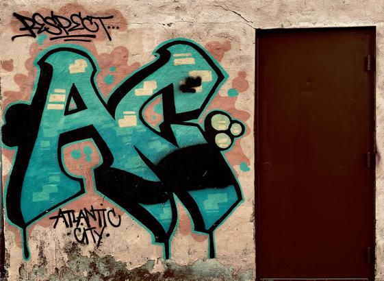A color, medium-angle photograph taken outside of a liquor store in Atlantic City, New Jersey. The focal point of the image is a vibrant piece of graffiti street art that is spray-painted onto a weathered, textured beige exterior wall next to a solid, dark brown metal door. The central focus is a large, stylized piece of lettering in a bright teal and turquoise hue, outlined in bold black. The letters "AC” feature blocky, geometric shapes with internal highlights of pale yellow and black shading to create a three-dimensional effect. Above the main letters, a black tag reads "RESPECT..." in a classic graffiti script with two underlines, while below the letters, "ATLANTIC CITY" is written in a more formal, sans-serif marker style. The wall itself shows signs of age, with peeling paint and dark moss atop water stains collecting along the base. The teal graffiti is set against a soft, cloud-like background of dusty rose spray paint that helps the letters pop from the neutral wall. The dark brown door on the right provides a stark, minimalist contrast to the colorful and chaotic energy of the artwork.