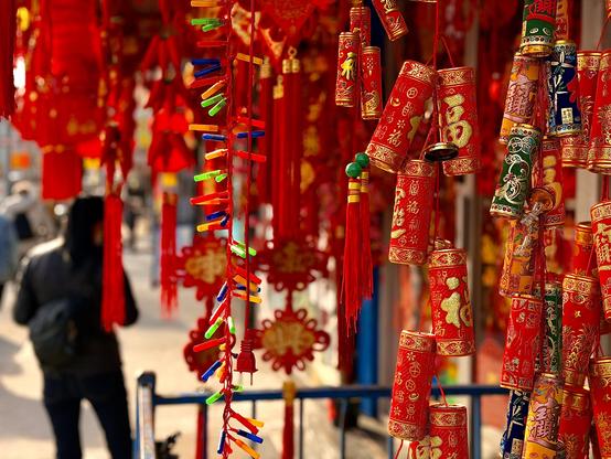 A color, medium-angle photograph of the street scene in the Bowery neighborhood of Manhattan. The focal point of the image is a storefront filled with Chinese new year decorations and string lights. In the mid-ground a person wearing black jeans, a black winter coat, and carrying a black crossbody bag are seen out of focus. In the distance are out of focus parked cars, buildings, and pedestrians. Morning sunlight is illuminating the scene.
