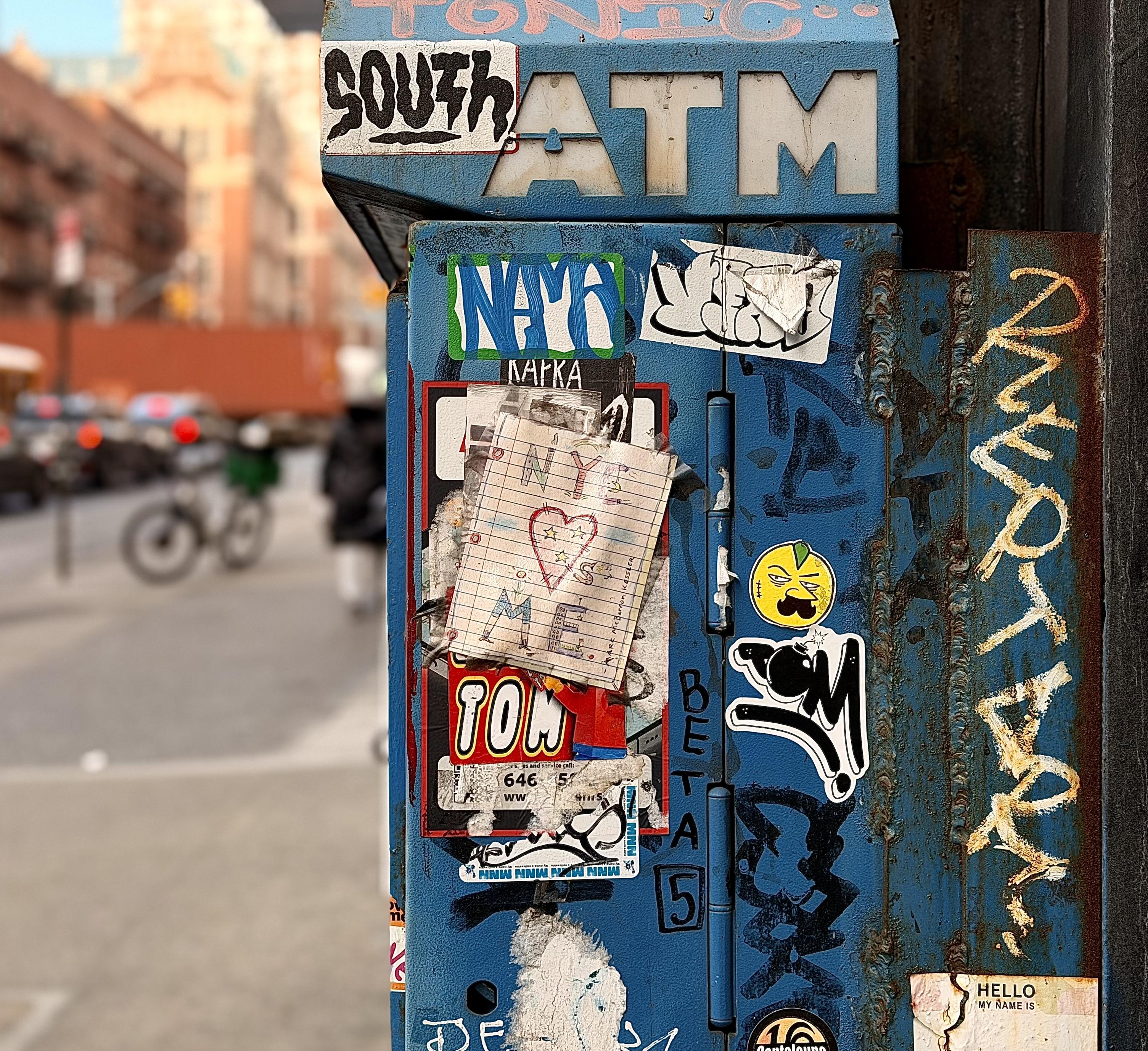 A color photograph in square aspect ratio. A close-up, shallow-depth-of-field shot focuses on the weathered side of a blue metal ATM kiosk along 10th Avenue in the Hell’s Kitchen neighborhood of Manhattan. The machine is heavily textured with layers of urban grit, rust, and street art. A prominent piece of lined notebook paper is taped to the center, featuring a hand-drawn "NYC ❤️ ME" doodle in colored markers. Surrounding this are various stickers, including a stylized "TOM" tag, a yellow smiley face with a mustache, and several black-and-white "slaps" with graffiti lettering. To the right, a rusted vertical metal beam shows a tall, hand-painted tag in weathered yellow paint. The background is softly blurred, showing the bustling environment of 10th Avenue during the day. A person in dark clothing walks away from the camera, and a bicycle is parked further down the sidewalk near a blurred street pole. In the distance, the warm tones of brick architecture and the soft red glow of car brake lights that are typical of a late afternoon light. The word "ATM" is visible in bold, white block letters at the top of the blue structure, partially obscured by a white and black "SOUTH" graffiti sticker. 