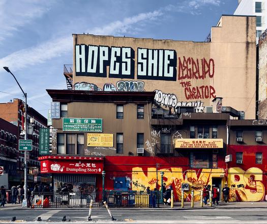 A color, wide-angle photograph in square aspect ratio. An eye-level capture of the vibrant street scene at the intersection of Chrystie and Grand Street in the Bowery neighborhood of Manhattan. The foreground is dominated by a weathered, two-story building featuring a red storefront for "Dumpling Story" on the left and a yellow facade on the right, both heavily covered in colorful graffiti, tags, and street art. Several people in winter clothing stand along the sidewalk or near the entrance of the Grand St subway station. In the upper left, a green street sign points toward "Chinatown Little Italy". The background is dominated by a large, beige multi-story building that serves as a massive canvas for urban art. Bold, blocky black-and-white graffiti reading "HOPES SHIE" spans the top, while large red dripping letters to the right read "DE GRUPO THE CREATOR." Numerous smaller tags and throw-ups in blue, white, and black ink fill the spaces between the windows and storefronts. The sky above is a bright, clear blue with thin, wispy white clouds.