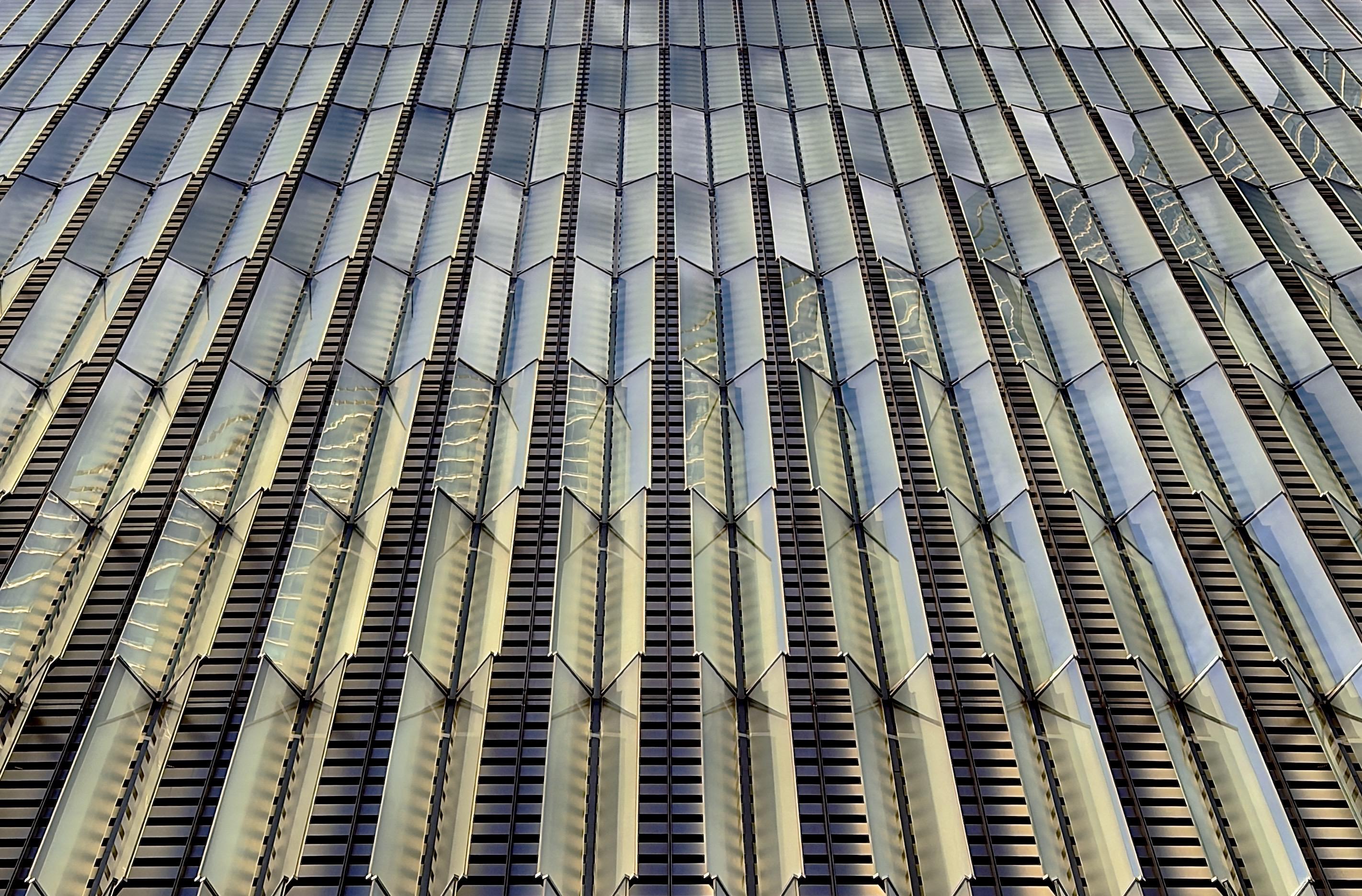 A color, medium-angle photograph in 5:4 orientation that captures the intricate architectural facade of the podium base at One World Trade Center in Manhattan on a mostly clear winter afternoon. The image is dominated by vertical columns of prismatic glass fins that project outward from the building’s surface in a rhythmic, staggered pattern. These triangular glass panels are set against a dark, recessed background of horizontal metal louvers, creating a high-contrast texture of light and shadow. The perspective is looking nearly straight up, causing the vertical lines to converge toward the top of the frame, emphasizing the immense scale and height of the structure. The glass fins are highly reflective, catching the ambient light to create a shimmering effect of silvers, soft golds, and pale blues. Some panels are angled in such a way that they reveal the structural metal ribs behind them, while others reflect the surrounding sky and neighboring buildings in distorted, geometric fragments. The interplay of the angled glass surfaces creates a bold and dynamic sense of depth, with the sharp edges of the fins slicing through the visual field. The overall aesthetic is one of modernity and precision, transforming a massive structural base into a delicate, light-catching sculpture of colorful brilliance.