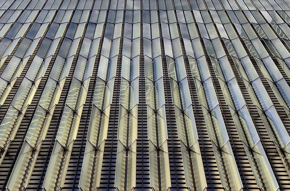 A color, medium-angle photograph in 5:4 orientation that captures the intricate architectural facade of the podium base at One World Trade Center in Manhattan on a mostly clear winter afternoon. The image is dominated by vertical columns of prismatic glass fins that project outward from the building’s surface in a rhythmic, staggered pattern. These triangular glass panels are set against a dark, recessed background of horizontal metal louvers, creating a high-contrast texture of light and shadow. The perspective is looking nearly straight up, causing the vertical lines to converge toward the top of the frame, emphasizing the immense scale and height of the structure. The glass fins are highly reflective, catching the ambient light to create a shimmering effect of silvers, soft golds, and pale blues. Some panels are angled in such a way that they reveal the structural metal ribs behind them, while others reflect the surrounding sky and neighboring buildings in distorted, geometric fragments. The interplay of the angled glass surfaces creates a bold and dynamic sense of depth, with the sharp edges of the fins slicing through the visual field. The overall aesthetic is one of modernity and precision, transforming a massive structural base into a delicate, light-catching sculpture of colorful brilliance.