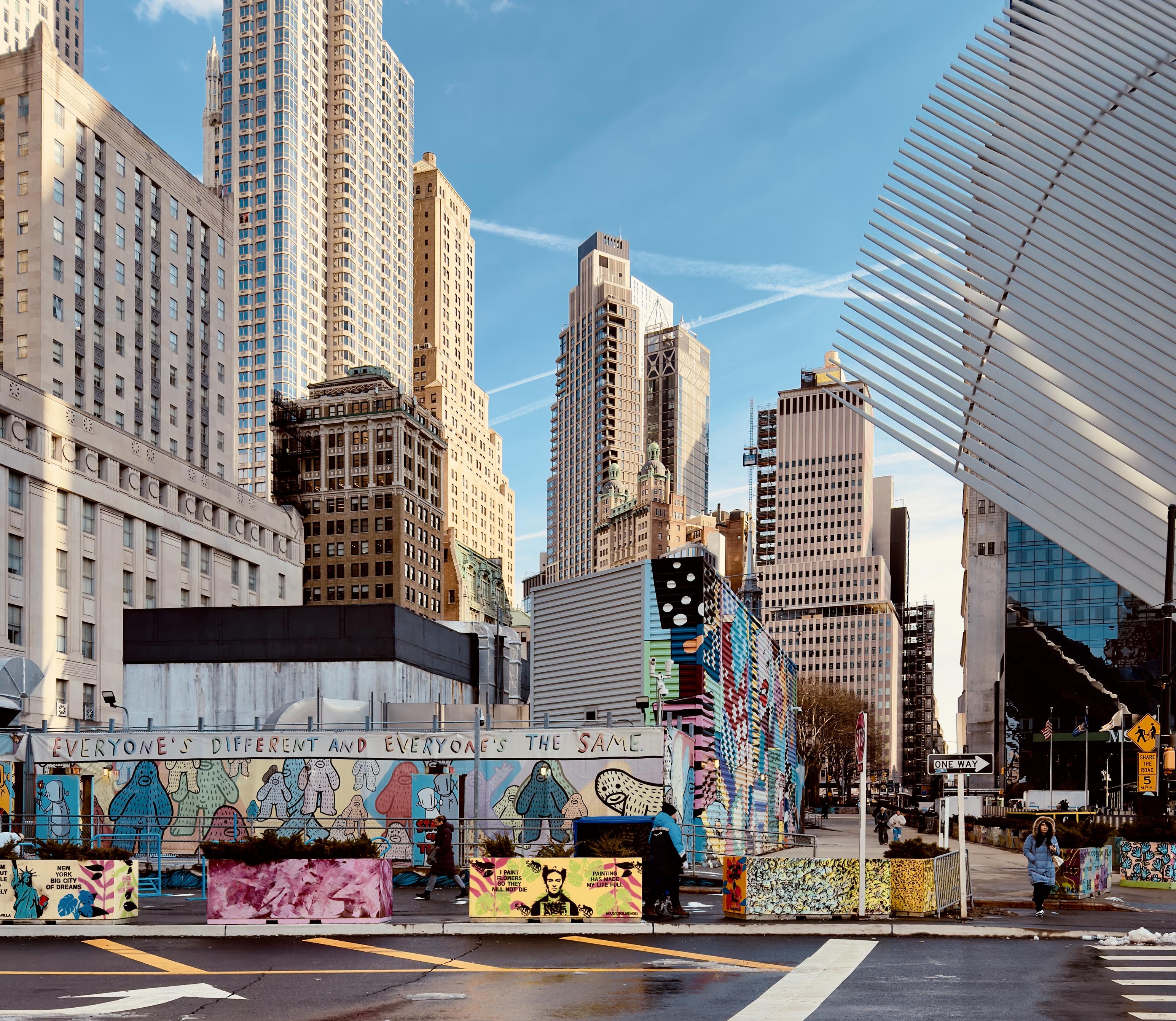 A color, wide-angle photograph in square aspect ratio captures the street scene outside of The Oculus, framed by the architecture of the World Trade Center site. In the foreground, a prominent mural by artist Brandon Sines features his signature character Frank Ape accompanied by the inclusive mantra, "EVERYONE'S DIFFERENT AND EVERYONE'S THE SAME." The mural stretches across several construction sheds, which are further decorated with smaller, eclectic art panels and pink-toned floral patterns. The scene is mostly still, but there are a few pedestrians walking and standing. The asphalt street is wet from winter precipitation earlier in the day. The background presents a striking contrast between modern glass skyscrapers and historic masonry. To the right, the distinct, white-ribbed "wings" of the Oculus transportation hub cut sharply into the sky. Behind the mural, a cluster of high-rise buildings includes the classic limestone facade of the Woolworth Building with its green copper roof, standing alongside contemporary residential towers and the sleek, dark glass of the newer World Trade Center structures. The composition is bathed in soft, late-afternoon sunlight, casting long shadows throughout the scene.