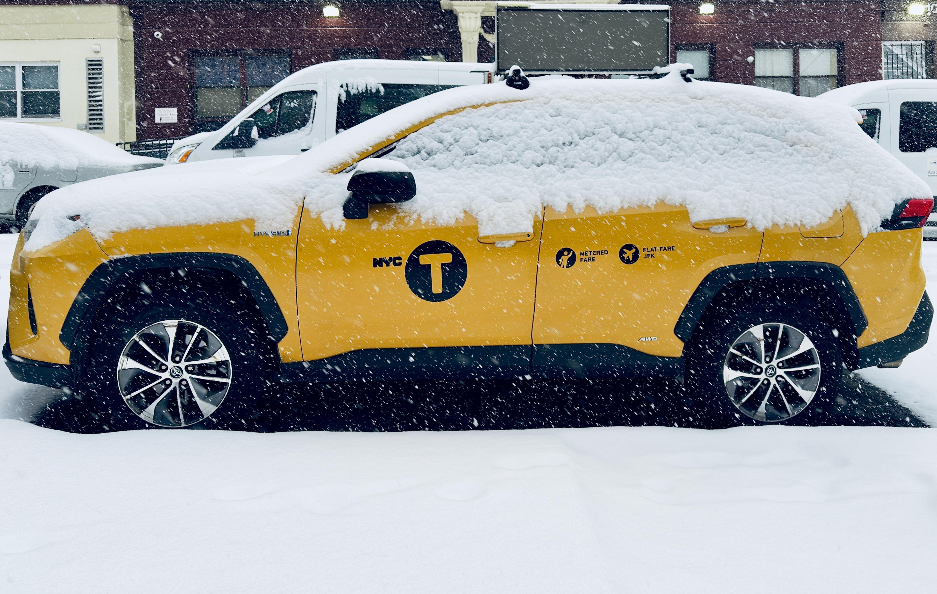 A color, medium-angle photograph in landscape orientation that captures a profile view of a vibrant yellow New York City taxi, specifically a modern SUV model, parked on a street during a snowstorm. The vehicle is heavily blanketed in a thick layer of fresh snow which covers the roof, the hood, and most of the side windows. The iconic black "NYC T" taxi emblem is clearly visible on the driver’s side door, alongside smaller black icons indicating metered fares and flat rates to JFK. Large, multi-spoke silver and black rims peek out from beneath the wheel wells, contrasted against the dark asphalt of the road and the accumulating snow on the ground. A digital advertisement board is mounted to the roof and turned off. The background is a hazy, urban setting obscured by thick, falling snowflakes. A white van is parked directly behind the taxi, and the dark brick facade of a building is behind the van.