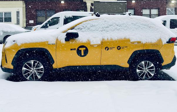 A color, medium-angle photograph in landscape orientation that captures a profile view of a vibrant yellow New York City taxi, specifically a modern SUV model, parked on a street during a snowstorm. The vehicle is heavily blanketed in a thick layer of fresh snow which covers the roof, the hood, and most of the side windows. The iconic black "NYC T" taxi emblem is clearly visible on the driver’s side door, alongside smaller black icons indicating metered fares and flat rates to JFK. Large, multi-spoke silver and black rims peek out from beneath the wheel wells, contrasted against the dark asphalt of the road and the accumulating snow on the ground. A digital advertisement board is mounted to the roof and turned off. The background is a hazy, urban setting obscured by thick, falling snowflakes. A white van is parked directly behind the taxi, and the dark brick facade of a building is behind the van.