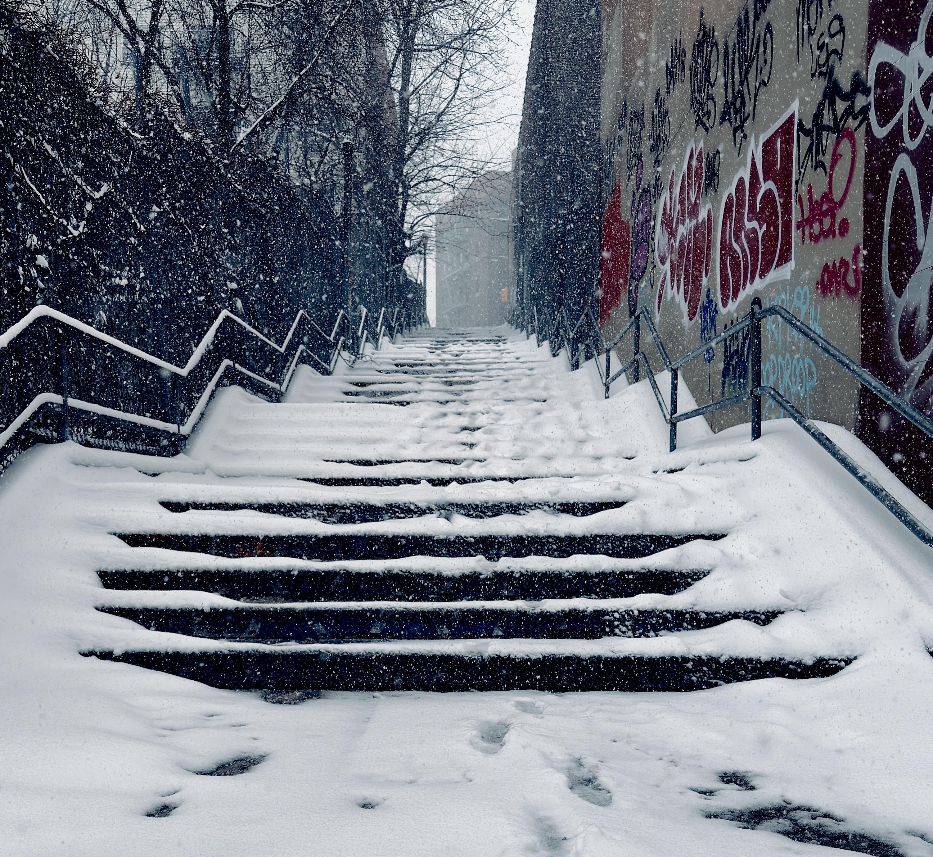A color, medium-angle photograph in square aspect ratio that captures a snow-covered outdoor stairwell in the Upper West Side neighborhood of Manhattan. Thick, fresh snow blankets the stone steps, though a narrow path in the center has been partially cleared by foot traffic and wind, revealing the dark treads beneath. To the right, a high concrete retaining wall is covered in vibrant red, white, and black graffiti, providing a sharp urban contrast to the soft white snow. On the left, a dark, leafless hedge and fence line runs parallel to the stairs. Metal handrails with a jagged, geometric design frame both sides of the staircase, disappearing upward into the heavy snowfall. The atmosphere is quiet and wintry, with large, visible snowflakes filling the air and softening the focus of the background. At the top of the long climb, the silhouette of a city building is faintly visible through the gray, overcast mist, emphasizing the steepness of the incline. Visibility is greatly reduced. The color palette is dominated by the cool blues and whites of the snow and the monochromatic tones of the city, punctuated only by the bold pops of color from the street art on the right.
