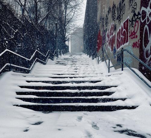 A color, medium-angle photograph in square aspect ratio that captures a snow-covered outdoor stairwell in the Upper West Side neighborhood of Manhattan. Thick, fresh snow blankets the stone steps, though a narrow path in the center has been partially cleared by foot traffic and wind, revealing the dark treads beneath. To the right, a high concrete retaining wall is covered in vibrant red, white, and black graffiti, providing a sharp urban contrast to the soft white snow. On the left, a dark, leafless hedge and fence line runs parallel to the stairs. Metal handrails with a jagged, geometric design frame both sides of the staircase, disappearing upward into the heavy snowfall. The atmosphere is quiet and wintry, with large, visible snowflakes filling the air and softening the focus of the background. At the top of the long climb, the silhouette of a city building is faintly visible through the gray, overcast mist, emphasizing the steepness of the incline. Visibility is greatly reduced. The color palette is dominated by the cool blues and whites of the snow and the monochromatic tones of the city, punctuated only by the bold pops of color from the street art on the right.