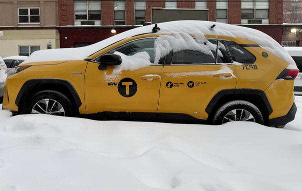 A color, medium-angle photograph in landscape orientation that captures an updated profile view of the bright yellow New York City taxi SUV parked on a street from the previous photo in this thread. The taxi is now partially buried after a significant amount of snowfall. The heavy, wind-sculpted snow from the peak remains piled high against the lower half of the vehicle, halfway obscuring the tires. A thick, white crust of snow still clings to the roof and hood, drooping over the tops of the windows in frozen, jagged edges. The iconic black "NYC T" medallion logo is centered on the front door, with the taxi’s identification number "7C98" visible toward the rear. The atmosphere has transitioned to a lighter snowfall, with smaller, more sparse flakes visible against the backdrop of a red brick building. The air appears clearer and the visibility has improved compared to earlier, revealing more detail in the surrounding urban environment, such as the windows of the building and a white van parked in the distance.