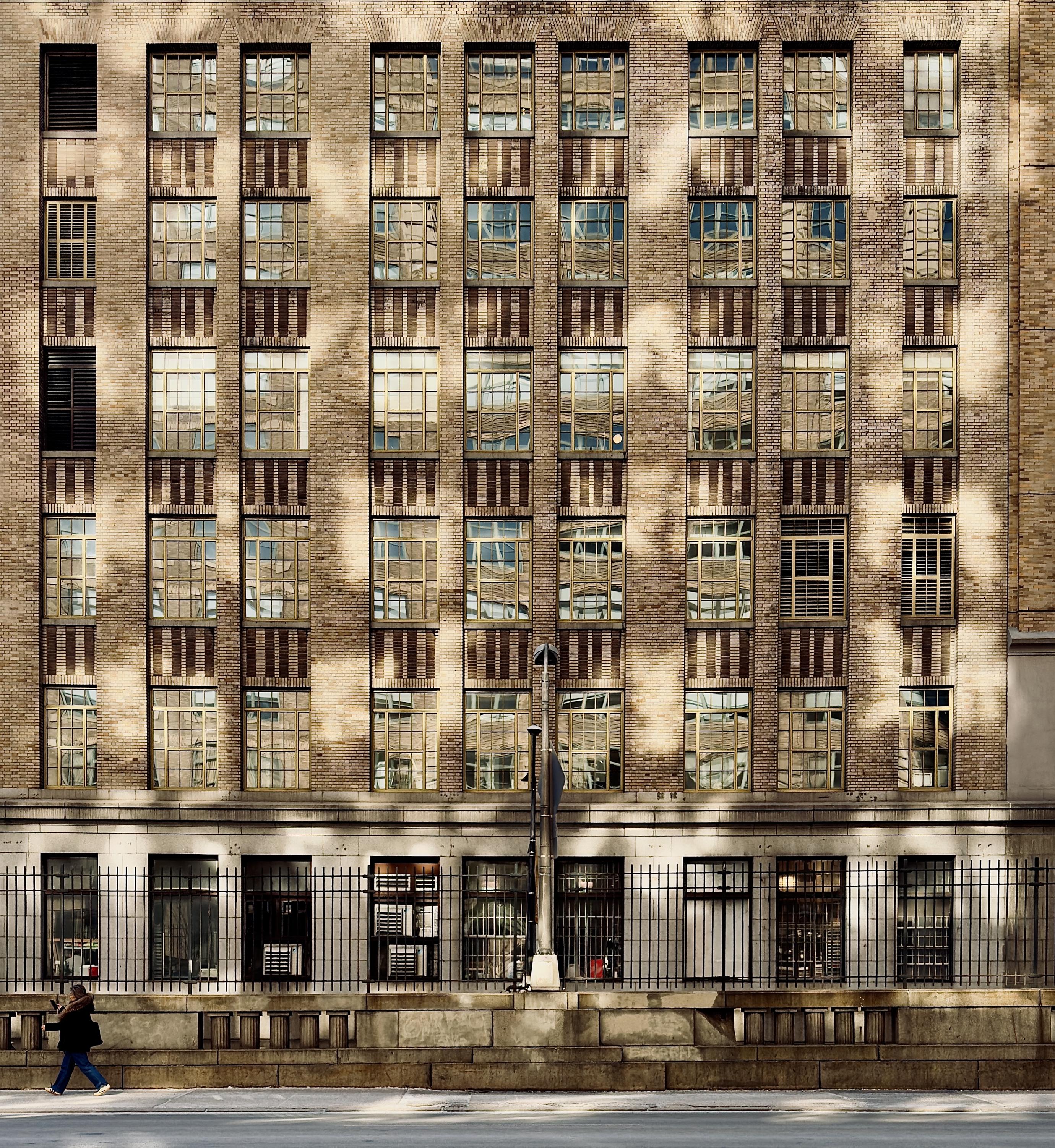 A color, medium-angle photograph in square aspect ratio that captures the textured facade of a six story Art Deco-style building in the Hudson Square neighborhood of Manhattan. The building is constructed from tan and brown brick with a rhythmic pattern of vertical window columns. The windows are framed by dark, decorative brickwork and set deep within the wall. Most of the glass panes are alive with brilliant, golden and blue-green reflections of nearby buildings and the brickwork has sunlight bouncing off from neighboring structure windows, creating distorted, painterly patches of light that dance across the building’s surface. On the ground level, a black wrought-iron fence runs the length of the building, partially obscuring the lower windows and a stone base. In the lower-left corner, a single pedestrian wearing a dark coat and blue jeans is walking briskly along the sidewalk near the left edge of the frame, with a phone in one hand. This helps provide a sense of scale to the massive structure. The lighting is late afternoon, as the long, dramatic shadows and high-contrast glares dominate the upper floors while the street level remains in cooler shade. A solitary street lamp stands centered in front of the building, its thin silhouette cutting through the middle of the frame against the complex, grid-like backdrop of the masonry.
