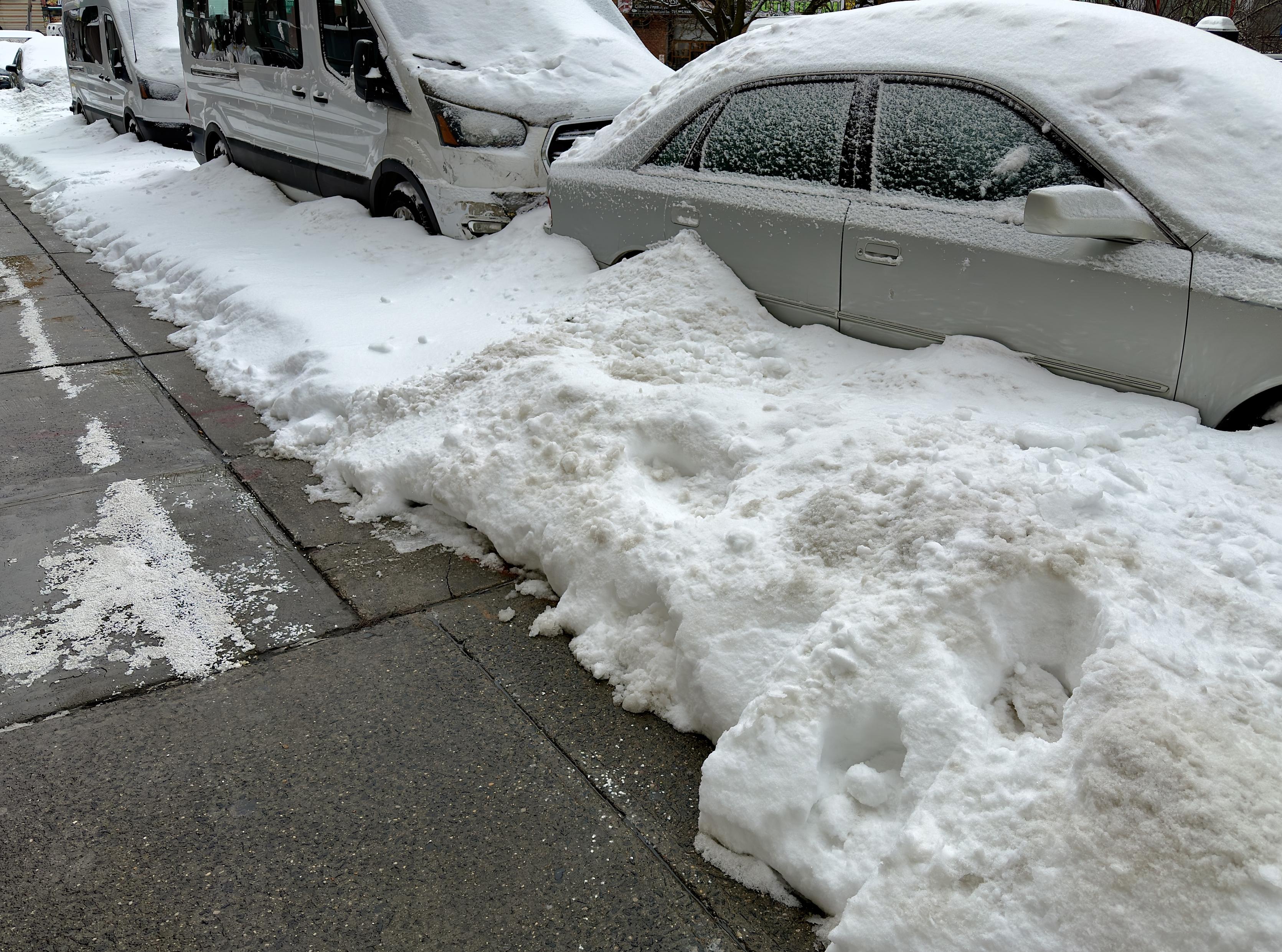 A color, wide-angle, eye-level photograph that captures a city sidewalk and a row of street-parked vehicles heavily buried under snow in the Upper West Side neighborhood of Manhattan. The left side of the frame features a clean, gray concrete sidewalk with visible cracks and a sparse dusting of white road salt. To the right of the walkway, a massive, continuous wall of dense, dirty-white snow has been plowed into a steep embankment. This snowbank completely obscures the lower halves of the vehicles, creating a jagged, uneven barrier that separates the clear pedestrian path from the street. The parked vehicles, including a light-colored sedan in the foreground and two large white transport vans further back, are draped in a thick, frozen blanket of snow. The sedan is nearly swallowed by the drifts, with snow piled high against its passenger doors and a dense layer covering its hood, roof, and windows. The vans behind it are similarly encased, their shapes softened by the heavy accumulation. The lighting is flat and overcast, casting soft shadows.