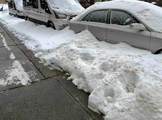 A color, wide-angle, eye-level photograph that captures a city sidewalk and a row of street-parked vehicles heavily buried under snow in the Upper West Side neighborhood of Manhattan. The left side of the frame features a clean, gray concrete sidewalk with visible cracks and a sparse dusting of white road salt. To the right of the walkway, a massive, continuous wall of dense, dirty-white snow has been plowed into a steep embankment. This snowbank completely obscures the lower halves of the vehicles, creating a jagged, uneven barrier that separates the clear pedestrian path from the street. The parked vehicles, including a light-colored sedan in the foreground and two large white transport vans further back, are draped in a thick, frozen blanket of snow. The sedan is nearly swallowed by the drifts, with snow piled high against its passenger doors and a dense layer covering its hood, roof, and windows. The vans behind it are similarly encased, their shapes softened by the heavy accumulation. The lighting is flat and overcast, casting soft shadows.