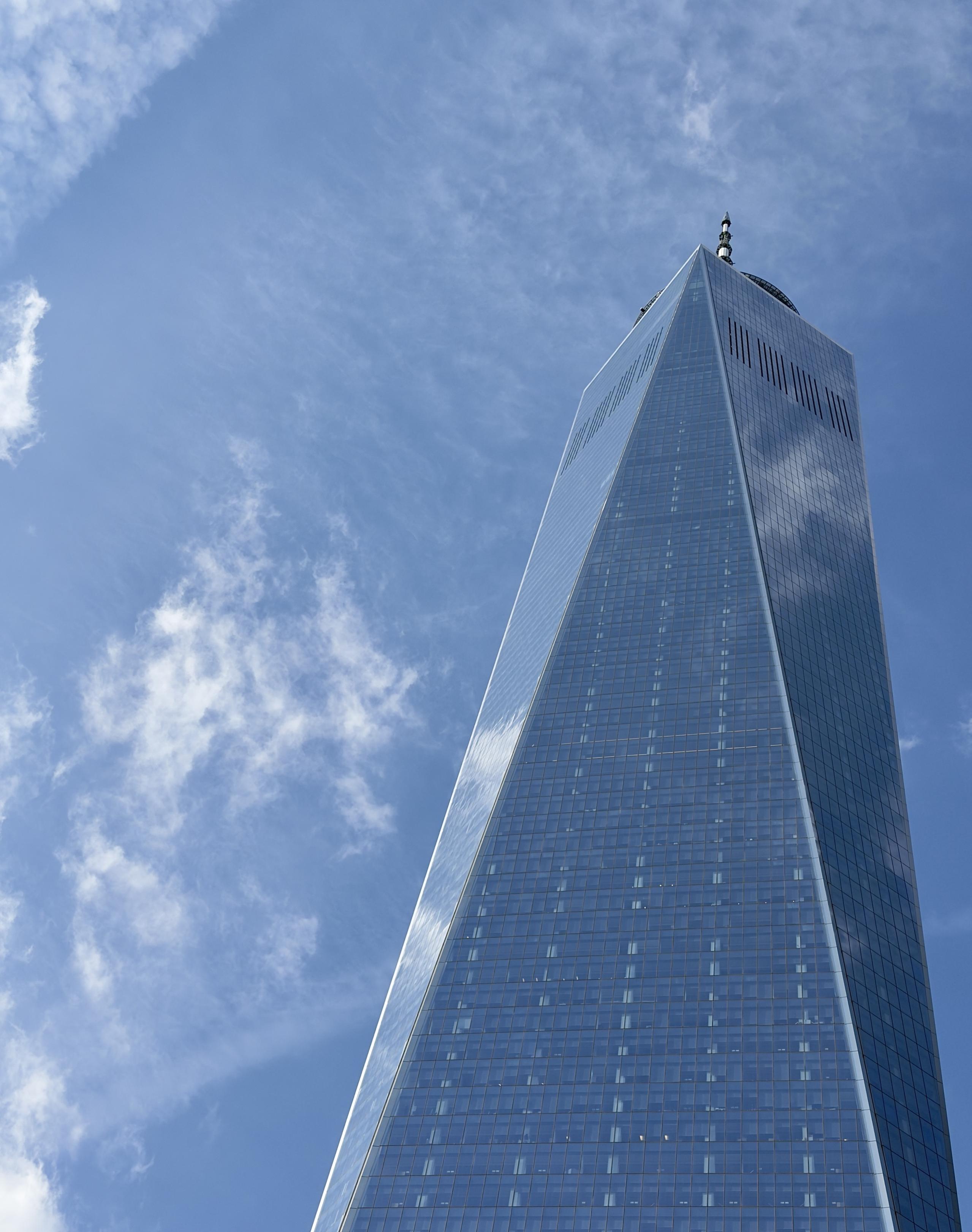 A color, low-angle photograph in portrait orientation that captures the One World Trade Center skyscraper in the Financial District of Manhattan. The tower is soaring upward against a vibrant, clear blue sky. The skyscraper's glass facade, composed of triangular planes, creates a shimmering, reflective surface that mirrors the passing wisps of white clouds. The perspective emphasizes the building’s immense height, making it appear to taper narrowly as it reaches toward the top of the frame, where its distinct communication spire pierces the bright atmosphere. The sky is a deep, crisp cerulean, dotted with soft, feathered white clouds. While there is no visible snow on the building’s slick, mirrored exterior, the intense sunlight hitting the glass facets creates a sharp contrast between the bright reflections and the deeper blue shadows on the shaded side of the tower. The overall composition is clean and minimalist, focusing on the geometric precision of the architecture and the renewed brilliance of the post-storm day.