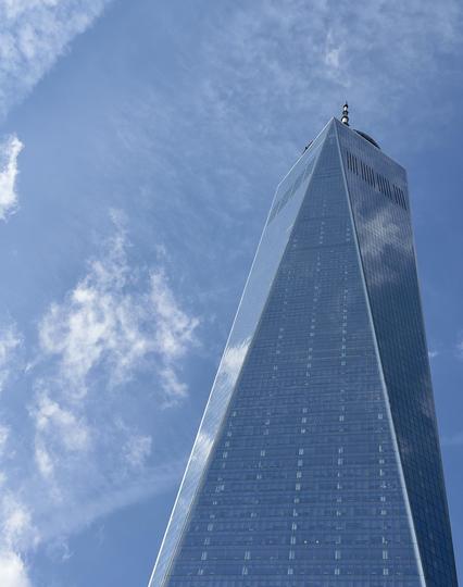A color, low-angle photograph in portrait orientation that captures the One World Trade Center skyscraper in the Financial District of Manhattan. The tower is soaring upward against a vibrant, clear blue sky. The skyscraper's glass facade, composed of triangular planes, creates a shimmering, reflective surface that mirrors the passing wisps of white clouds. The perspective emphasizes the building’s immense height, making it appear to taper narrowly as it reaches toward the top of the frame, where its distinct communication spire pierces the bright atmosphere. The sky is a deep, crisp cerulean, dotted with soft, feathered white clouds. While there is no visible snow on the building’s slick, mirrored exterior, the intense sunlight hitting the glass facets creates a sharp contrast between the bright reflections and the deeper blue shadows on the shaded side of the tower. The overall composition is clean and minimalist, focusing on the geometric precision of the architecture and the renewed brilliance of the post-storm day.