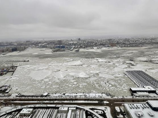 A color, wide-angle photograph taken from an elevated position that captures a view looking west across the Hudson River from Manhattan on a frigid, overcast day. The river is heavily congested with large, jagged ice floes and slush, covering nearly the entire surface of the water except for a dark, narrow channel near the New Jersey shoreline. In the foreground, the snow-dusted rooftops of Manhattan, including industrial buildings and a transit yard, lead toward the riverbank. Several piers extend into the icy water, including a large, modern rectangular pier on the right and a construction area with cranes on the left. The horizon is dominated by the New Jersey skyline, featuring a dense expanse of low-rise buildings and industrial structures blanketed in white snow. The sky is a heavy, uniform slate gray, blending into the pale, frozen landscape and muting the colors of the city. A few small boats are visible, navigating the treacherous icy patches, while the distant shoreline stretches out under the hazy, wintry atmosphere.