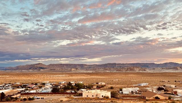 A color, wide-angle photograph in landscape orientation that captures a serene sunrise over the desert community of Greenehaven, Arizona from an elevated view. In the foreground, scattered residential homes with flat roofs and earthy, southwestern architecture sit nestled among sparse desert vegetation. The middle ground transitions into a vast, arid plateau of golden-brown sand and scrub brush, leading the eye toward a dramatic horizon of flat-topped mesas and rugged canyon walls. The rock formations are layered in shades of tan and pale cream, glowing softly under the early morning light. The expansive sky dominates the upper two-thirds of the image, filled with a dense layer of textured altocumulus clouds. These clouds are illuminated with a soft palette of pastel pinks, pale oranges, and hints of lavender where the sun crests the horizon, contrasting against patches of pale blue and muted grey. The overall atmosphere is peaceful and vast, emphasizing the quiet beauty of the high desert.