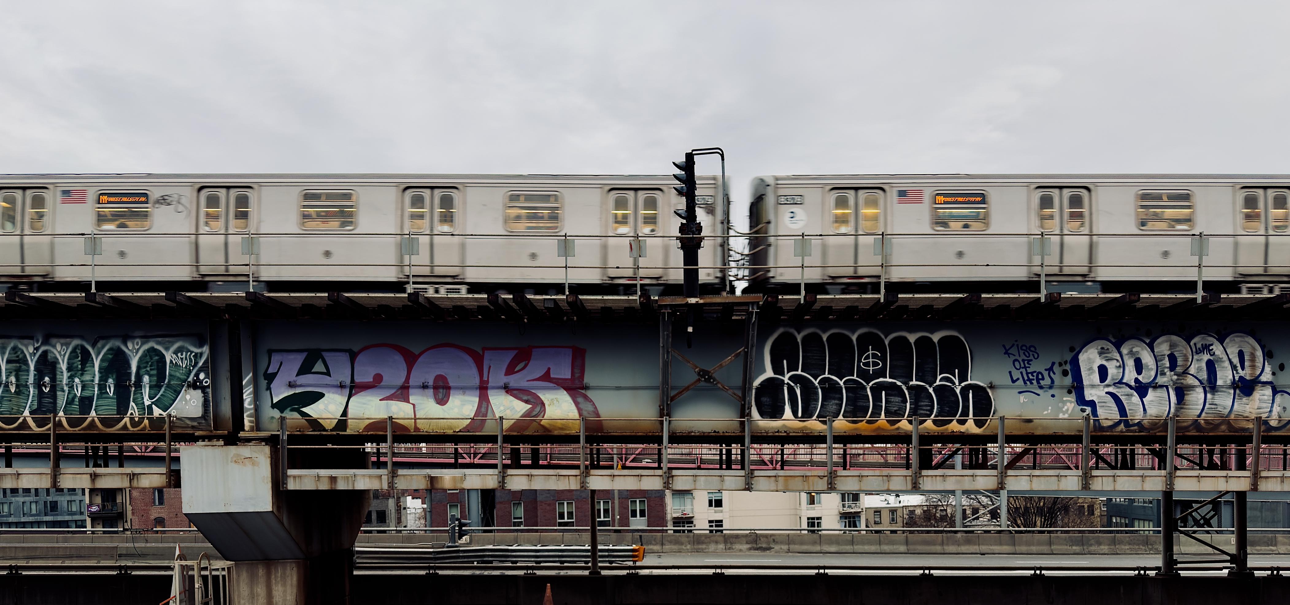 A color, wide-angle photograph in landscape orientation that captures a silver New York City M train in motion as it traverses an elevated track along the Williamsburg Bridge in Brooklyn. The train cars, adorned with American flag decals and glowing yellow interior lights, appear slightly blurred, conveying a sense of speed against a pale, overcast afternoon winter sky. Below the tracks, the thick steel support beams of the bridge structure are heavily covered in vibrant, large-scale graffiti throw-ups in shades of blue, purple, and black. A vertical black signal post stands prominently between the two visible train cars, cutting through the horizontal lines of the composition. In the lower third of the frame, the industrial landscape of the Williamsburg area is visible beneath the bridge’s infrastructure. A secondary metal walkway with a safety railing runs parallel to the tracks, while the brick facades of apartment buildings and a glimpse of the pinkish-red suspension cables of the Williamsburg Bridge peek through the background. The overall atmosphere is gritty and quintessentially urban.
