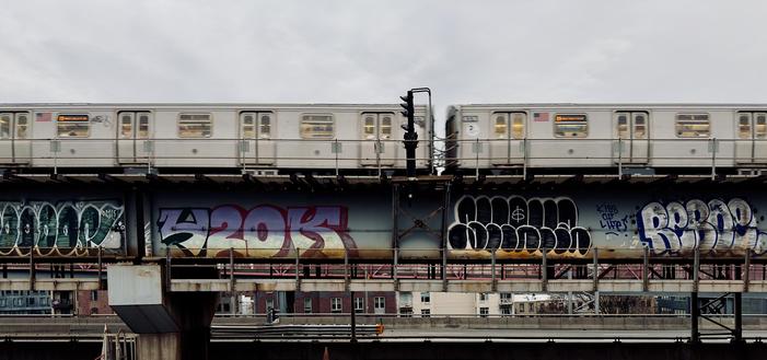 A color, wide-angle photograph in landscape orientation that captures a silver New York City M train in motion as it traverses an elevated track along the Williamsburg Bridge in Brooklyn. The train cars, adorned with American flag decals and glowing yellow interior lights, appear slightly blurred, conveying a sense of speed against a pale, overcast afternoon winter sky. Below the tracks, the thick steel support beams of the bridge structure are heavily covered in vibrant, large-scale graffiti throw-ups in shades of blue, purple, and black. A vertical black signal post stands prominently between the two visible train cars, cutting through the horizontal lines of the composition. In the lower third of the frame, the industrial landscape of the Williamsburg area is visible beneath the bridge’s infrastructure. A secondary metal walkway with a safety railing runs parallel to the tracks, while the brick facades of apartment buildings and a glimpse of the pinkish-red suspension cables of the Williamsburg Bridge peek through the background. The overall atmosphere is gritty and quintessentially urban.