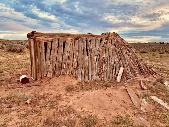 A color, medium-angle photograph in 5:4 aspect ratio that captures the weathered framework of an unfinished traditional Navajo hogan, set against the vast, open landscape of the high desert. The structure is composed of dozens of thick, vertically aligned juniper logs, leaning inward to form an oval shape. These logs are rough-hewn and silvery-brown from sun exposure, with many still retaining their natural bark texture and knots. On the left side, a rectangular entryway is framed by flat wooden planks. The ground immediately surrounding the hogan is a mix of fine, reddish-orange sand and sparse, low-lying desert scrub, with several discarded wooden planks and a rusted metal bucket scattered nearby. The background transitions into an expansive horizon of rolling, arid plains dotted with sagebrush and small shrubs under a beautifully expressive sky. The sky is filled with a dense layer of streaky, blue-gray altocumulus clouds that catch the soft, golden light of the setting sun, creating a pale yellow glow along the horizon. This lighting casts soft shadows across the undulating desert floor and highlights the rugged, textured surfaces of the timber.