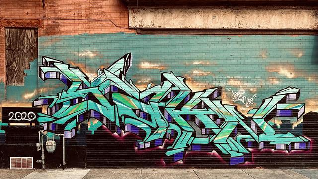 A color, wide-angle, eye-level photograph in landscape orientation that captures a vibrant piece of wildstyle graffiti spanning the length of a teal-painted brick wall in Atlantic City. The mural features intricate, interlocking geometric letters in shades of mint green, turquoise, and emerald, accented with sharp white highlights and deep purple shadows that create a three-dimensional effect. The piece is set against a background of soft, glowing orange and yellow patches that mimic a sunset, while the bottom of the mural is grounded by a dark black "cityscape" silhouette with a glowing magenta aura. To the left, the year "2020" is painted in white lettering on a black block. The mural is situated on the side of a weathered building, where the upper portion consists of traditional red-orange bricks and a weathered, off-white cornice. To the far left, a doorway is boarded up with dark plywood, partially covered by the teal paint of the mural's background. At the base of the wall, a gray concrete sidewalk runs parallel to the building, featuring a silver gas meter and various utility pipes. The overall lighting is warm and cinematic, emphasizing the contrast between the gritty textures of the city street and the polished, energetic artistry of the graffiti.