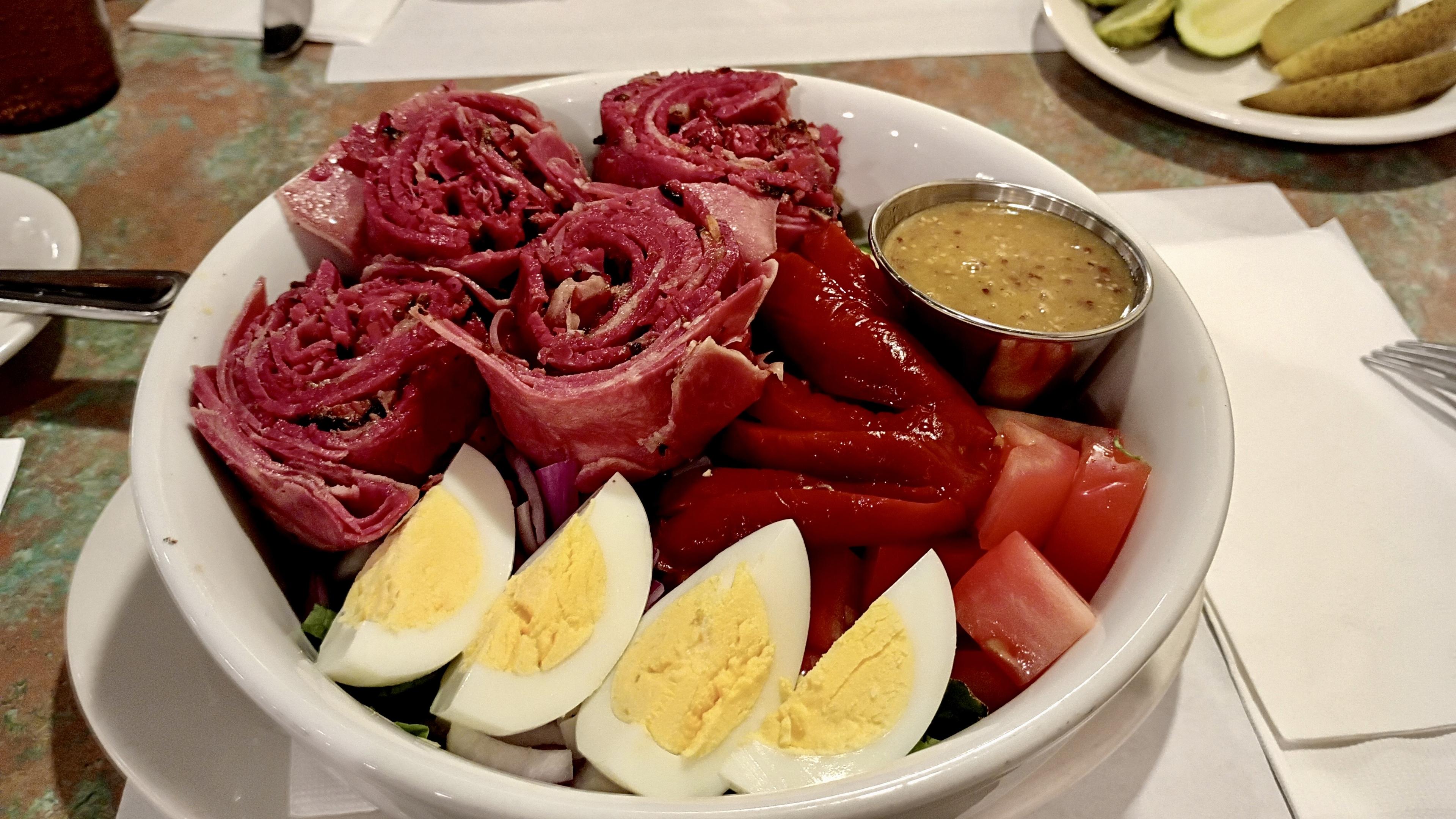 A color, medium-angle photograph in landscape orientation taken at a diner table in Liebman’s Deli in the Riverdale neighborhood of the Bronx. A white ceramic bowl is filled with a delicatessen chef salad, prominently featuring four large, rose-like swirls of thinly sliced, bright red pastrami arranged across the top. To the right of the meat, several thick strips of roasted red peppers add a deep crimson hue, accompanied by a small stainless steel ramekin filled with grainy white wine yellow mustard. The front of the bowl displays four hard-boiled egg quarters with pale yellow yolks, leaning against a bed of fresh greens, sliced red onions, and chunks of red tomato. The salad sits on a white plate atop a vintage-style teal and brown speckled Formica tabletop. In the background, a white side plate holds traditional deli accompaniments, including bright green pickled tomatoes and several half-sour pickle spears. A white paper napkin and a stainless steel fork are visible to the right, while the corner of a beverage glass containing iced tea, and a silver spoon rest to the left.