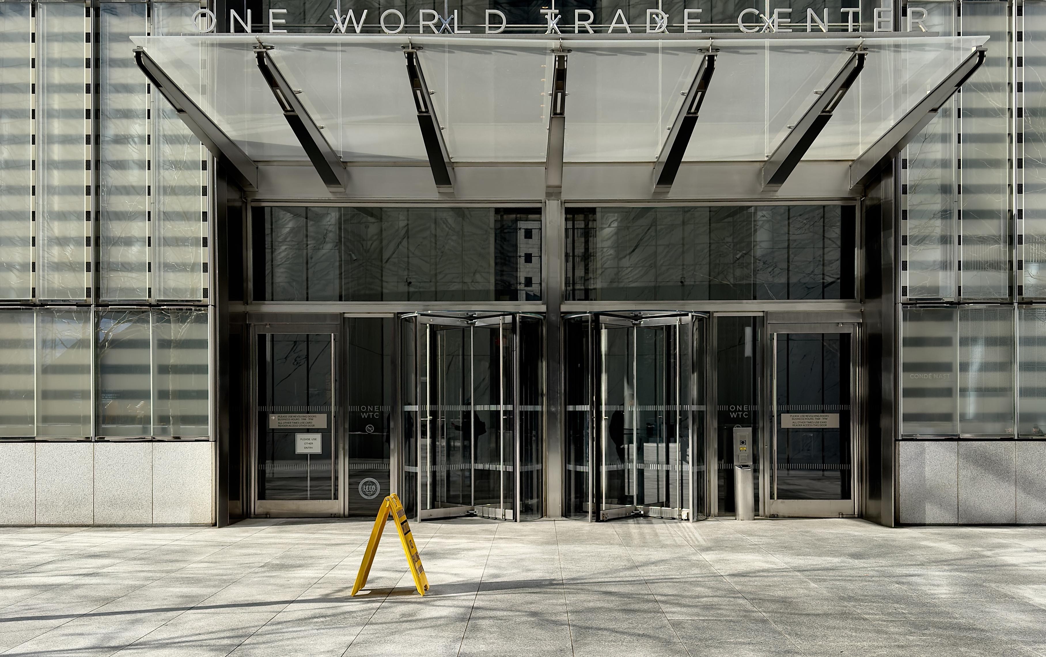 A color, wide-angle photograph in landscape orientation that captures the modern ground-floor entrance of One World Trade Center in the Financial District neighborhood of Manhattan. The composition is centered on two sets of large glass revolving doors, flanked by standard glass entry doors. Above the entrance, a massive, angular glass and metal canopy extends outward, supported by thick steel beams. The words "ONE WORLD TRADE CENTER" are displayed in clean, silver sans-serif metal lettering across the top of the canopy's frame. The facade is characterized by tall, vertical glass panels with subtle horizontal frosted stripes, creating a sleek and reflective architectural texture. The ground in the foreground is made of light grey stone pavers, illuminated by bright, direct sunlight that casts sharp shadows from a nearby leafless tree. A single bright yellow "Caution" A-frame sign stands prominently on the pavement in front of the left revolving door, adding a pop of color to the otherwise monochromatic silver and grey palette. Through the transparency of the glass doors, the interior lobby appears dimly lit, with faint reflections of the surrounding urban environment visible on the polished exterior surfaces. To the far right, a small "Condé Nast" sign is visible on the glass, indicating a primary tenant of the building.