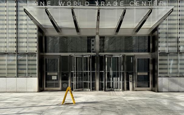 A color, wide-angle photograph in landscape orientation that captures the modern ground-floor entrance of One World Trade Center in the Financial District neighborhood of Manhattan. The composition is centered on two sets of large glass revolving doors, flanked by standard glass entry doors. Above the entrance, a massive, angular glass and metal canopy extends outward, supported by thick steel beams. The words "ONE WORLD TRADE CENTER" are displayed in clean, silver sans-serif metal lettering across the top of the canopy's frame. The facade is characterized by tall, vertical glass panels with subtle horizontal frosted stripes, creating a sleek and reflective architectural texture. The ground in the foreground is made of light grey stone pavers, illuminated by bright, direct sunlight that casts sharp shadows from a nearby leafless tree. A single bright yellow "Caution" A-frame sign stands prominently on the pavement in front of the left revolving door, adding a pop of color to the otherwise monochromatic silver and grey palette. Through the transparency of the glass doors, the interior lobby appears dimly lit, with faint reflections of the surrounding urban environment visible on the polished exterior surfaces. To the far right, a small "Condé Nast" sign is visible on the glass, indicating a primary tenant of the building.