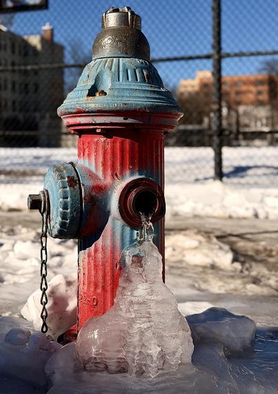 A color, medium-angle photograph in portrait orientation of the winter street scene in the Harlem neighborhood of Manhattan. The majority of the image is a vibrant fire hydrant that stands in the foreground, heavily weathered with chipped red and light blue paint. It is positioned on the ice and snow-covered ground against a backdrop of a chain-link fence and blurred city buildings under a clear blue sky. A steady, slow leak from one of its side ports has frozen into a large, translucent ice formation that cascades down to the ground, creating a thick, jagged mound of ice at its base. The hydrant itself shows signs of age, with rusted metal edges and a heavy metal chain hanging from a capped nozzle on the left side. The lighting is bright and crisp, casting a distinct shadow of the hydrant across the surrounding slush and snow. The ice formation is the focal point of the image, glistening with frozen ripples and textures that capture the sunlight. The contrast between the industrial, colorful metal of the hydrant and the organic, frozen shape of the water creates a cold, winter atmosphere. In the distance, the soft-focus urban setting shows a fenced in basketball court covered uniformly in snow.