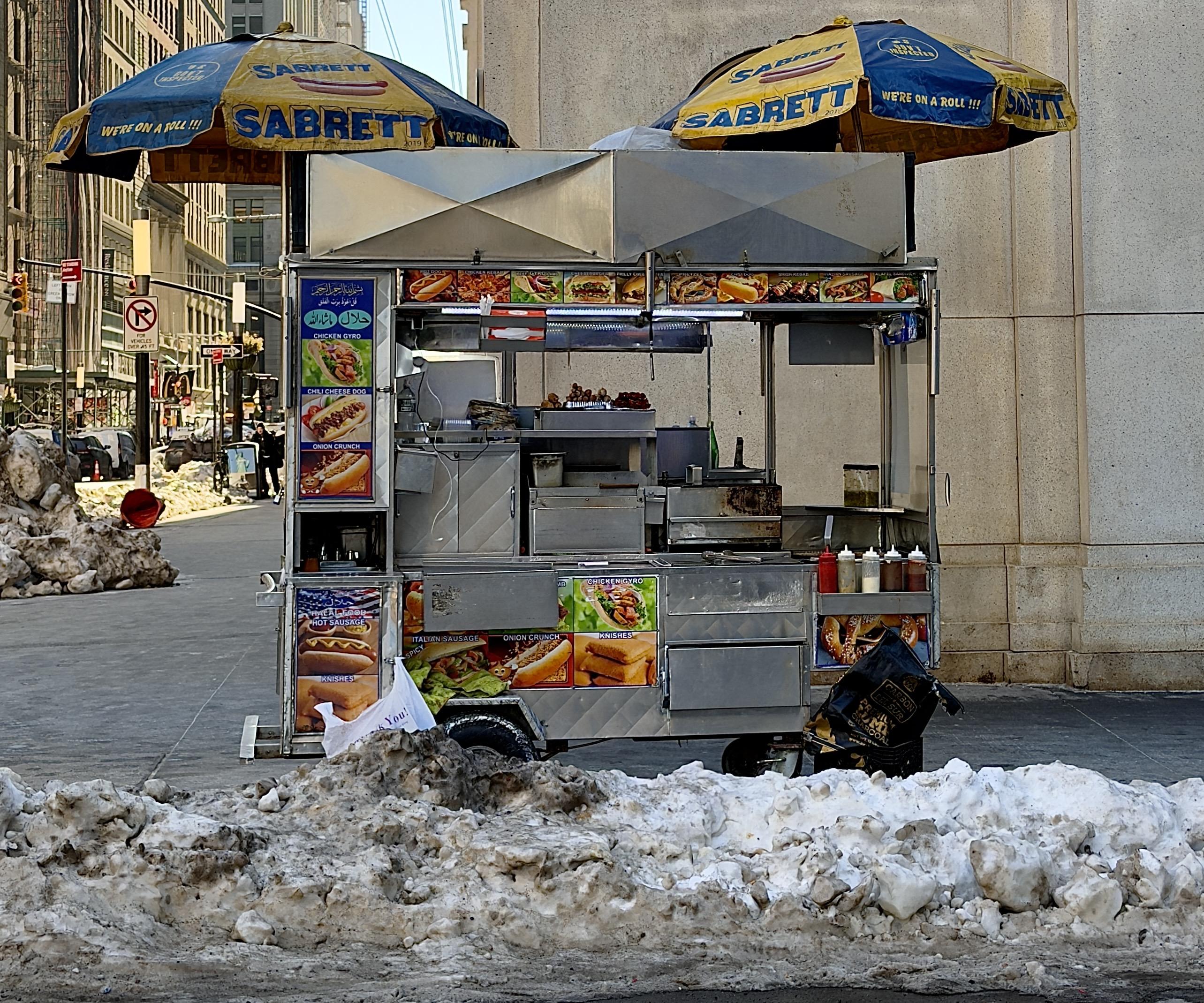 A color, wide-angle photograph in square aspect ratio that captures a classic silver metal street food cart stationed on a sidewalk in the Financial District neighborhood of Manhattan. The scene is framed by large mounds of dirty snow in the foreground. The cart is sheltered by two iconic blue and yellow Sabrett umbrellas featuring the slogan "We’re on a Roll!!!" and illustrations of hot dogs. The cart’s exterior is densely decorated with colorful photographic menus showcasing a variety of offerings, including halal chicken gyros, chili cheese dogs, Italian sausages, and soft pretzels. Several condiment squeeze bottles and a small pile of knishes are visible on the service counter, while a black bag sits on the lower ledge. The background reveals the streetscape, characterized by imposing stone buildings and a glimpse of a busy intersection. A "No Standing" sign and a traffic light are visible to the left, while a few pedestrians in winter clothing walk along the distant sidewalk. The lighting is bright and crisp, casting soft shadows on a cold winter afternoon.