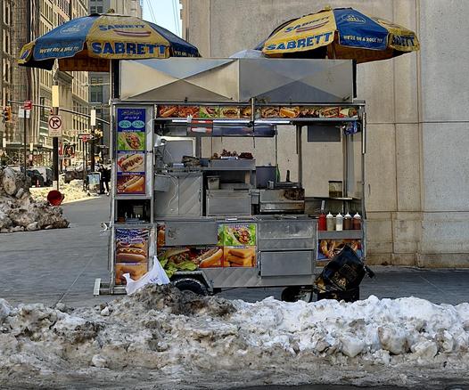 A color, wide-angle photograph in square aspect ratio that captures a classic silver metal street food cart stationed on a sidewalk in the Financial District neighborhood of Manhattan. The scene is framed by large mounds of dirty snow in the foreground. The cart is sheltered by two iconic blue and yellow Sabrett umbrellas featuring the slogan "We’re on a Roll!!!" and illustrations of hot dogs. The cart’s exterior is densely decorated with colorful photographic menus showcasing a variety of offerings, including halal chicken gyros, chili cheese dogs, Italian sausages, and soft pretzels. Several condiment squeeze bottles and a small pile of knishes are visible on the service counter, while a black bag sits on the lower ledge. The background reveals the streetscape, characterized by imposing stone buildings and a glimpse of a busy intersection. A "No Standing" sign and a traffic light are visible to the left, while a few pedestrians in winter clothing walk along the distant sidewalk. The lighting is bright and crisp, casting soft shadows on a cold winter afternoon.