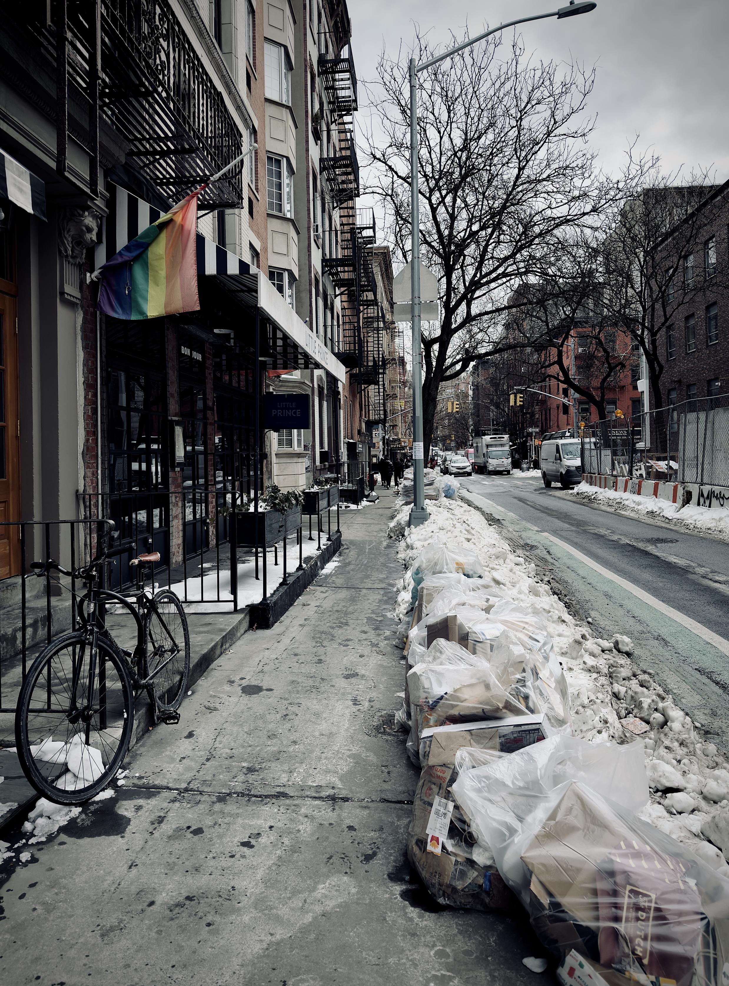 A color, wide-angle photograph in portrait orientation that captures the winter time street scene along Prince Street in the SoHo neighborhood of Manhattan. A bicycle is leaned against a metal railing in front of "Little Prince," a restaurant with a striped blue-and-white awning and a prominent rainbow Pride flag hanging half-curled above the entrance. Patches of white snow cling to the bike tires and the base of the building. The architecture features red brick facades, black metal fire escapes climbing up the multi-story buildings, and a decorative stone lion head carving near the restaurant door. On the right side of the frame, the narrow street is lined with large piles of dirty snow and a long row of clear plastic trash bags filled with cardboard and paper recycling awaiting collection. A green-painted bike lane runs parallel to the sidewalk, separated by the slushy snowbanks. In the distance, several white delivery trucks and vans are parked or moving down the street facing the camera, framed by the bare, dark branches of a deciduous tree, a lamppost, and more red brick buildings. The overcast morning light is dramatically lighting the scene evenly with no shadows.