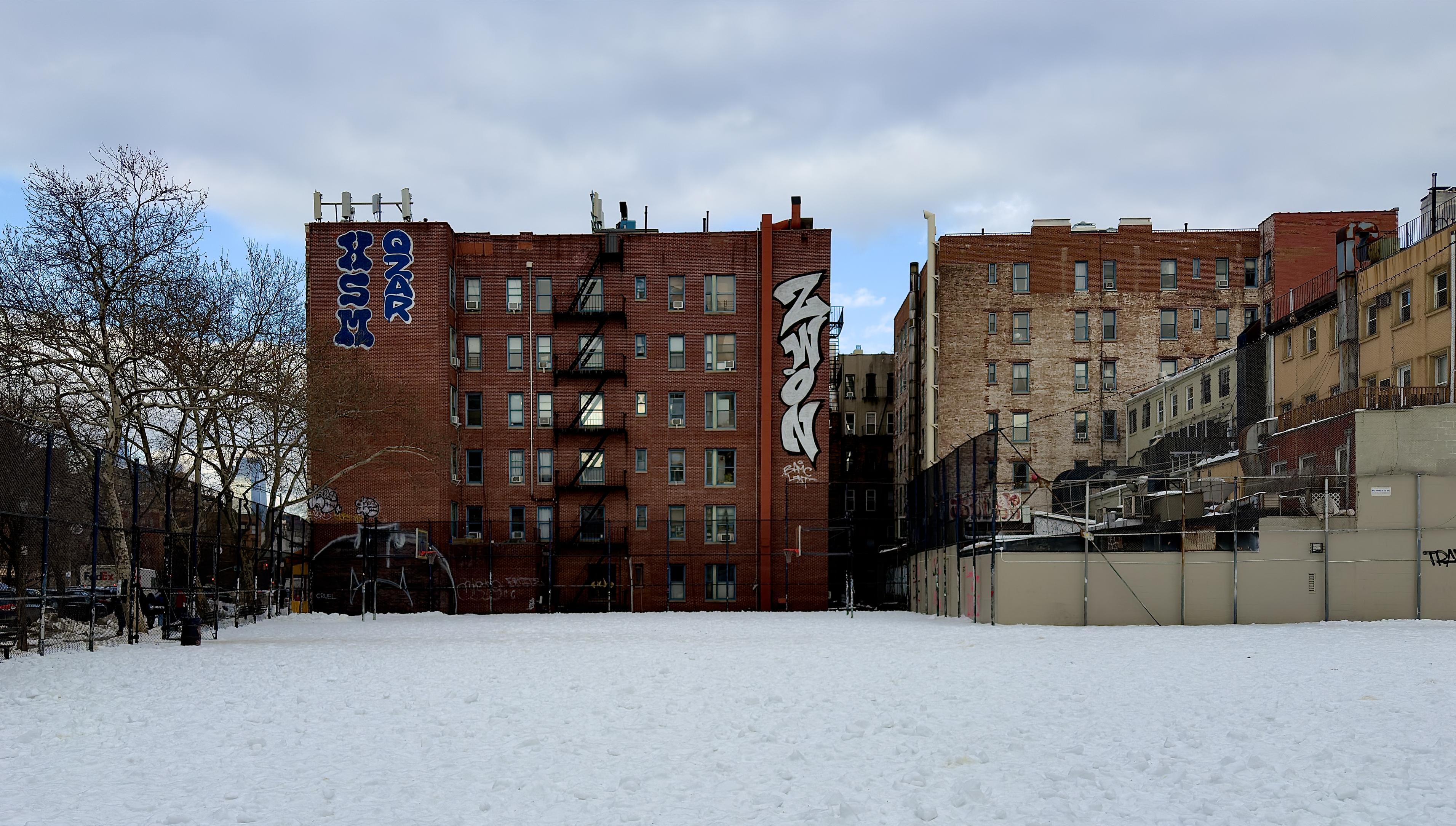 A color, wide-angle photograph in landscape orientation that captures the street scene along Houston St in the Greenwich Village neighborhood of Manhattan. The foreground is filled with a snow-covered ballpark. The afternoon winter sky is most cloudy, with small patches of light blue shining through. The ballpark is a flat expanse of bright white snow, textured with small mounds and footprints. A dark chain-link fence runs along the left and back of the field, with a basketball hoop visible near the center-left. Bare, wintry trees with intricate branch networks stand behind the fence on the left, while a few figures, parked cars, and a FedEx truck are faintly visible in the distance beyond the park's perimeter. The background is defined by several multi-story brick apartment buildings. The central building is a prominent six-story structure of dark red brick featuring a black metal fire escape zigzagging down its facade. Very large, stylized graffiti tags are painted high on its side walls: blue and white lettering on the left and vertical white and black block letters on the right. To its right, an older, light-colored brick building shows significant weathering and exposed patches of beige stone. A lower, beige-colored structure with a corrugated metal roof and various HVAC units sits in the lower right foreground, separated from the snowy field by a high fence.