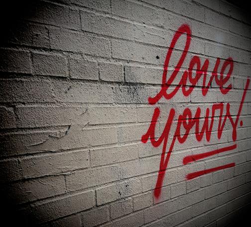 A color, medium-angle, close-up photograph in square aspect ratio that features a textured white brick wall with red spray-painted graffiti that reads "love yours!" in a flowing, cursive script. The text is positioned on the right side of the frame, underlined by two bold, horizontal red streaks, and ends with a prominent exclamation point. The wall’s surface shows aged details like small cracks, dark scuff marks, and uneven mortar lines, all captured with a shallow depth of field that causes the left side of the image to softly blur. A heavy black vignette circles the edges of the frame, focusing the viewer’s eye on the vibrant red lettering.