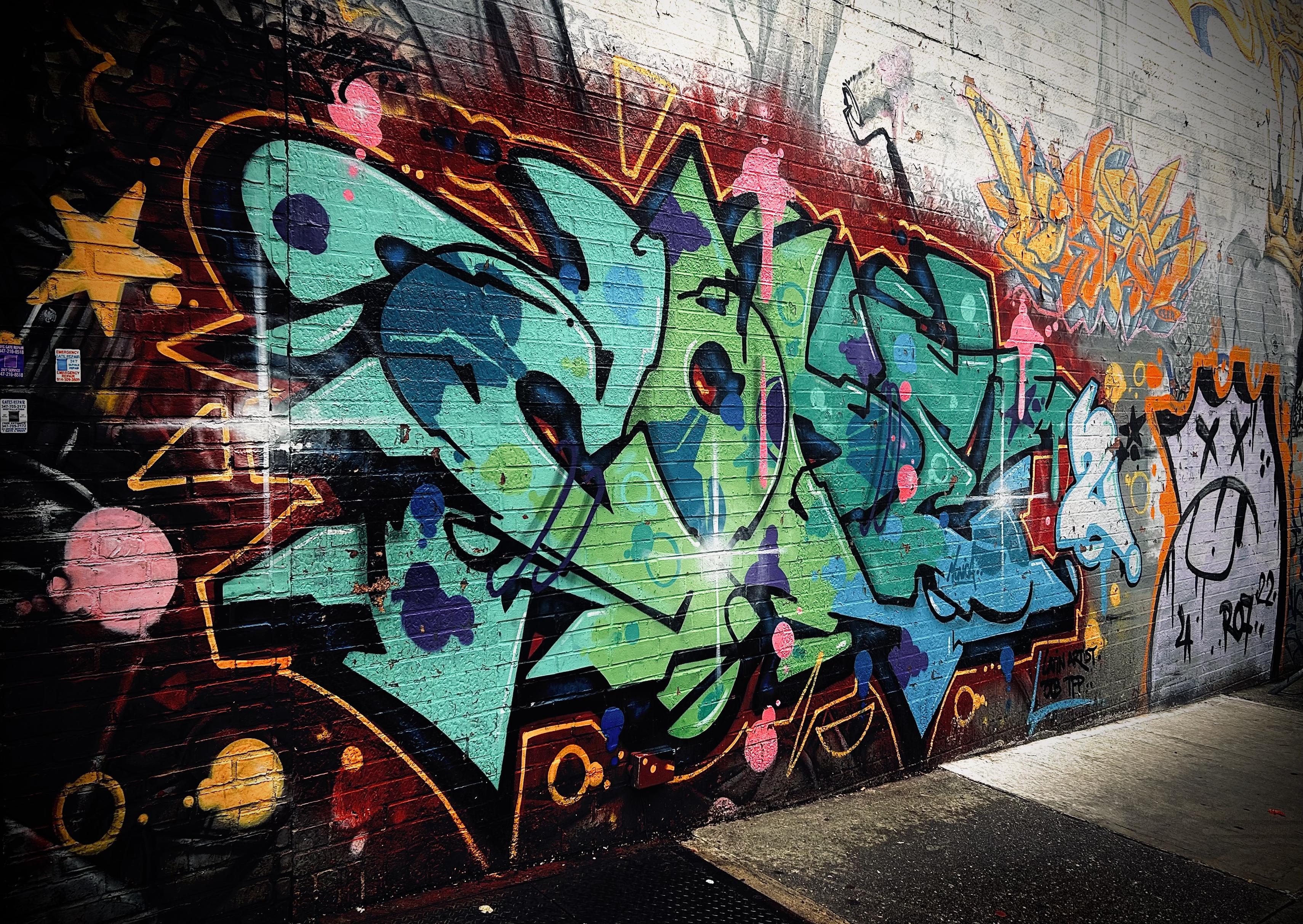 A color, wide-angle photograph in landscape orientation that captures a vibrant wild style graffiti mural painted on a textured brick wall in an alleyway in the Bronx. The central piece features large, interlocking letters in shades of teal, lime green, and cerulean blue, outlined with bold black and accented by a thin, glowing yellow-orange border. The letters are stylized with sharp, aggressive points and dynamic directional arrows, overlaid with splatters of pink and purple paint and circular "bubble" patterns. White "shine" marks or stars are placed strategically on the letter edges to simulate a reflective, three-dimensional metallic effect against the darker background of the brick. To the right of the main piece, a smaller, simplified orange and yellow tag sits above a black-and-white illustration of a character with "X" eyes and a crown. Faded remnants of older tags and star shapes in yellow and pink are scattered across the left side of the frame. The lighting is moody and dramatic, with heavy vignetting that darkens the corners of the image, drawing focus to the brightly colored center. The ground consists of a dark, damp pavement sidewalk in the lower right. Morning sunlight evenly illuminates the scene.