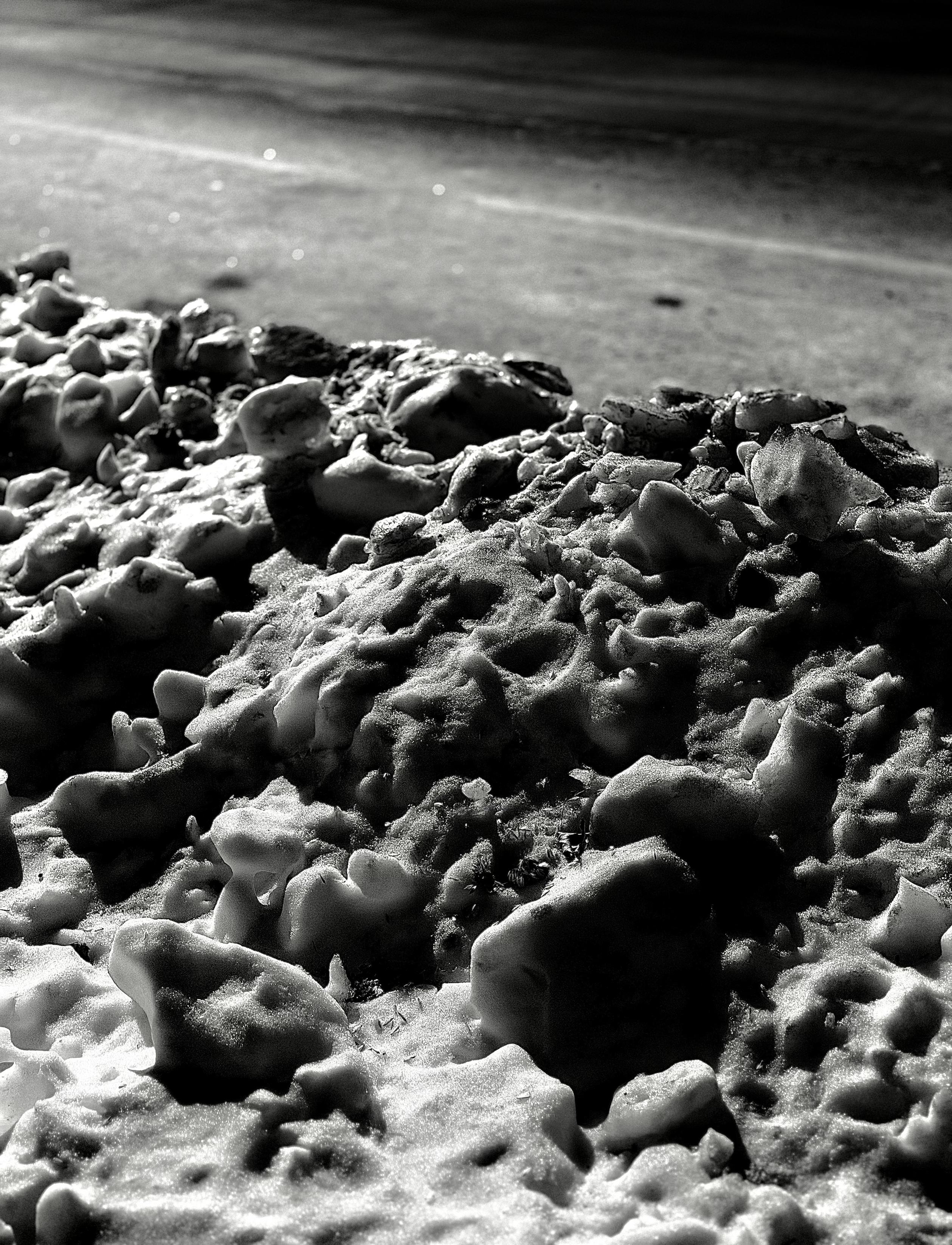 A noir, medium-angle photograph in portrait orientation that captures a pile of dirty snow banked along the edge of a street in the upper west side neighborhood of Manhattan. It is nighttime, and the scene is illuminated by a street lamp outside the frame. The foreground is a chaotic landscape of clumpy ice chunks and gritty slush, pitted with small craters and dusted with dark soot that emphasizes its rough, uneven texture. Harsh light from the upper left casts deep, dramatic shadows across the mounds, giving the frozen mass a mountain-like appearance. In the background, the smooth, dark asphalt of the road stretches away in a soft blur, with faint glints of light reflecting off the pavement.