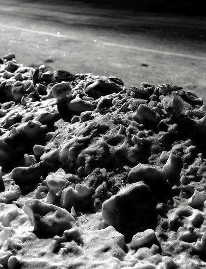 A noir, medium-angle photograph in portrait orientation that captures a pile of dirty snow banked along the edge of a street in the upper west side neighborhood of Manhattan. It is nighttime, and the scene is illuminated by a street lamp outside the frame. The foreground is a chaotic landscape of clumpy ice chunks and gritty slush, pitted with small craters and dusted with dark soot that emphasizes its rough, uneven texture. Harsh light from the upper left casts deep, dramatic shadows across the mounds, giving the frozen mass a mountain-like appearance. In the background, the smooth, dark asphalt of the road stretches away in a soft blur, with faint glints of light reflecting off the pavement.