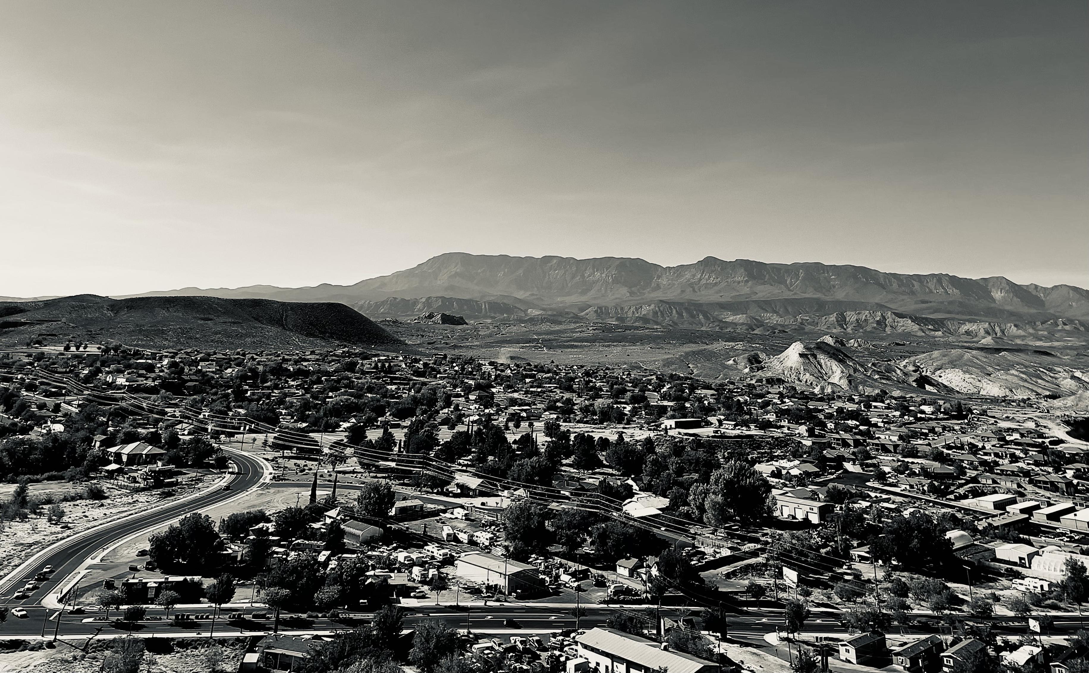 A noir, wide-angle photograph in landscape orientation. A high-angle view that captures the sprawling residential and commercial landscape of Hurricane, Utah, set against a dramatic desert backdrop. In the foreground and midground, a dense network of suburban streets, houses, and small industrial buildings is interspersed with mature trees and desert vegetation. A prominent curved multi-lane road sweeps through the lower-left side of the frame, leading the eye toward the heart of the town. The overall texture of the town reflects a blend of developed neighborhoods and rugged, open terrain, with various structures casting soft shadows under a clear, bright sky. The background is dominated by a vast mountain range with jagged peaks and layered ridgelines that stretch across the entire horizon. To the right, unique geological formations—including a distinctive white-capped conical hill—rise from the valley floor.