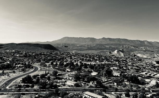 A noir, wide-angle photograph in landscape orientation. A high-angle view that captures the sprawling residential and commercial landscape of Hurricane, Utah, set against a dramatic desert backdrop. In the foreground and midground, a dense network of suburban streets, houses, and small industrial buildings is interspersed with mature trees and desert vegetation. A prominent curved multi-lane road sweeps through the lower-left side of the frame, leading the eye toward the heart of the town. The overall texture of the town reflects a blend of developed neighborhoods and rugged, open terrain, with various structures casting soft shadows under a clear, bright sky. The background is dominated by a vast mountain range with jagged peaks and layered ridgelines that stretch across the entire horizon. To the right, unique geological formations—including a distinctive white-capped conical hill—rise from the valley floor.